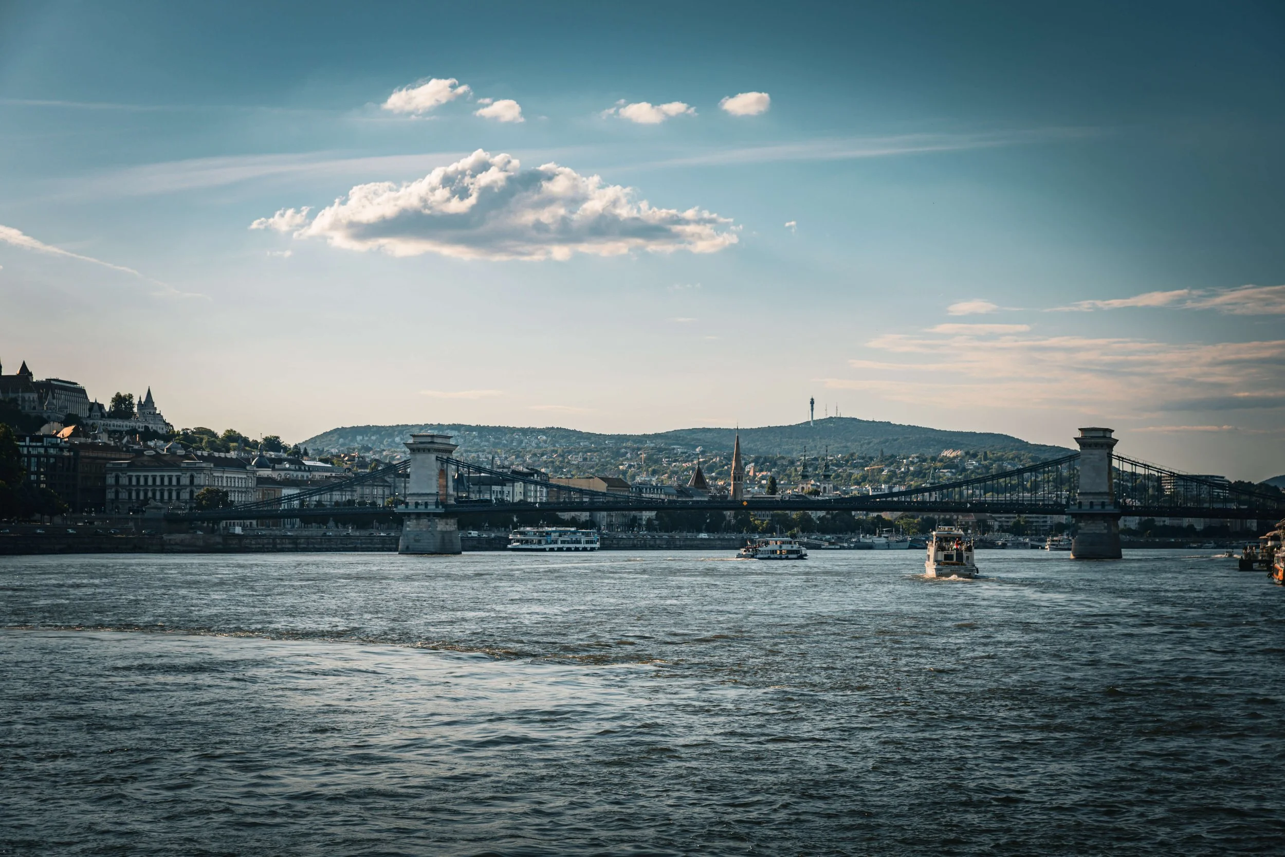 View of a river with boats, a bridge, city buildings, and a hill with a tower in the background under a partly cloudy sky.