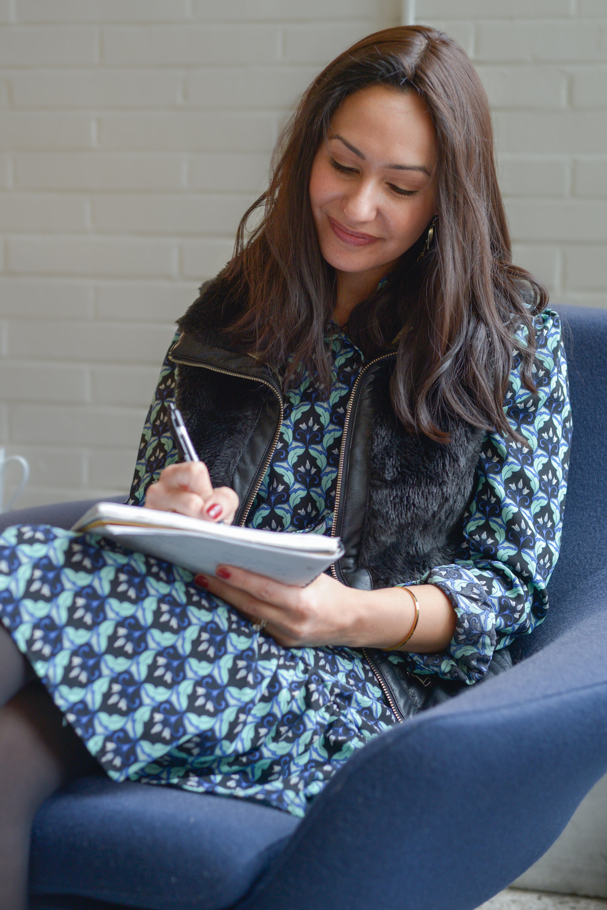 Rebecca is sitting on a blue chair, writing in a notebook with a pen, wearing a dress with a blue floral pattern and a black vest.
