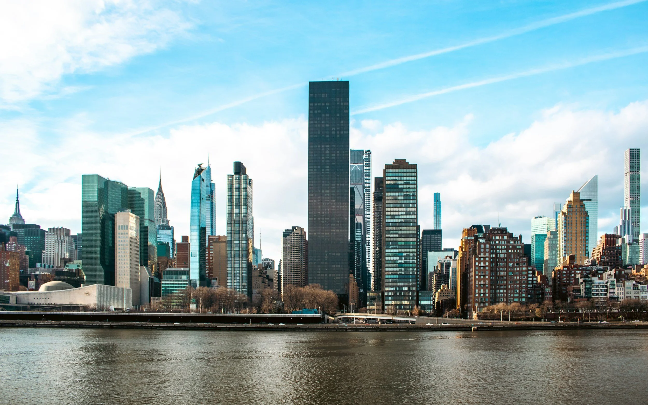 View of Manhattan skyline with tall buildings and skyscrapers along the water, under blue sky with clouds.