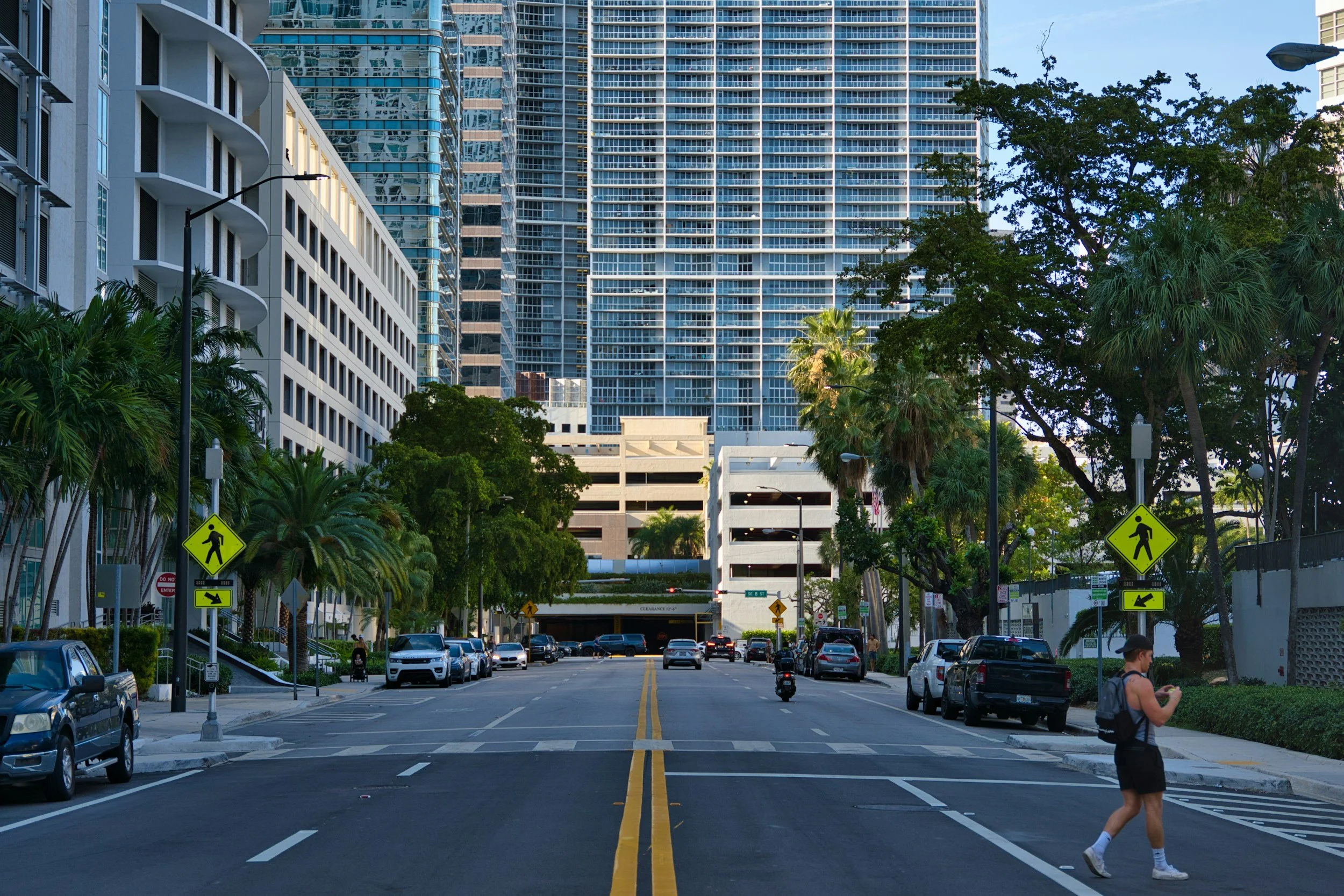 An urban street scene with tall modern glass buildings, trees lining the street, and a man crossing the street on a pedestrian crosswalk while looking at his phone, cars parked along the sides, and signage indicating pedestrian crossing.