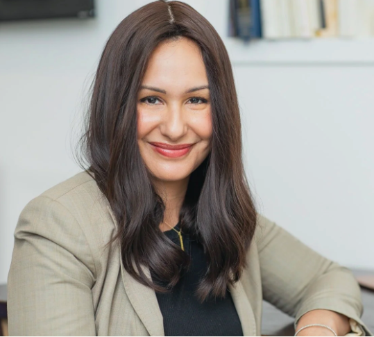 Rebecca Marcus, LCSW, smiling, dressed in a beige blazer and black top, sitting in an office setting with bookshelves in the background.