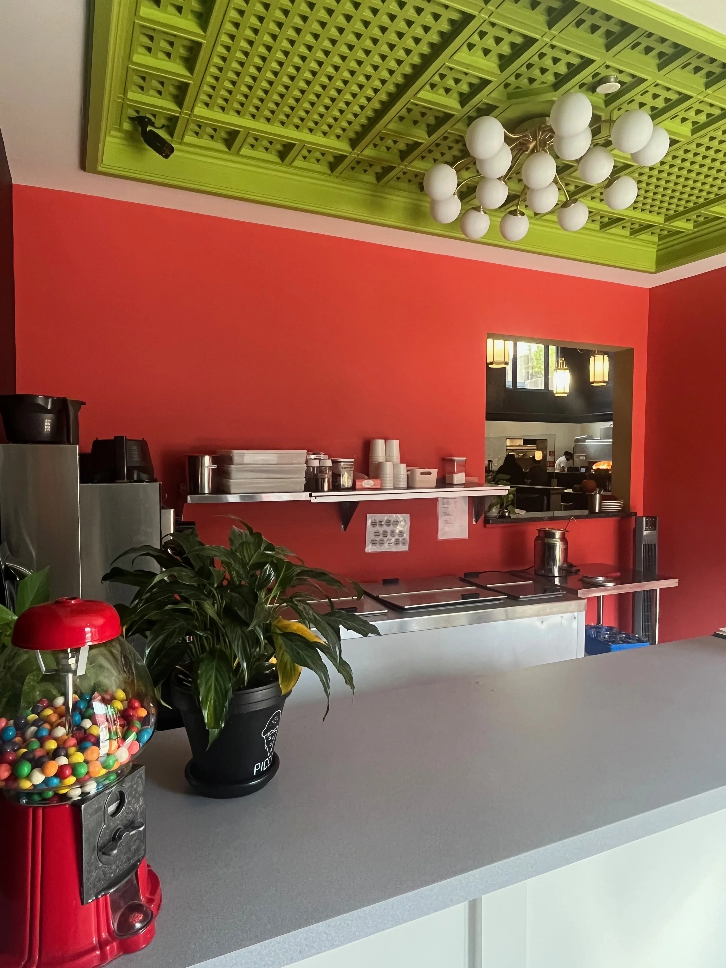 A gray counter with a red gumball machine filled with colorful gumballs, a potted plant, and a black ice cream cone cup with white lettering in the foreground. In the background, a vibrant red wall with a green ceiling and a white spherical chandelier hanging from above. Part of a kitchen or restaurant is visible through a window in the wall, with two hanging lamps and a person working.