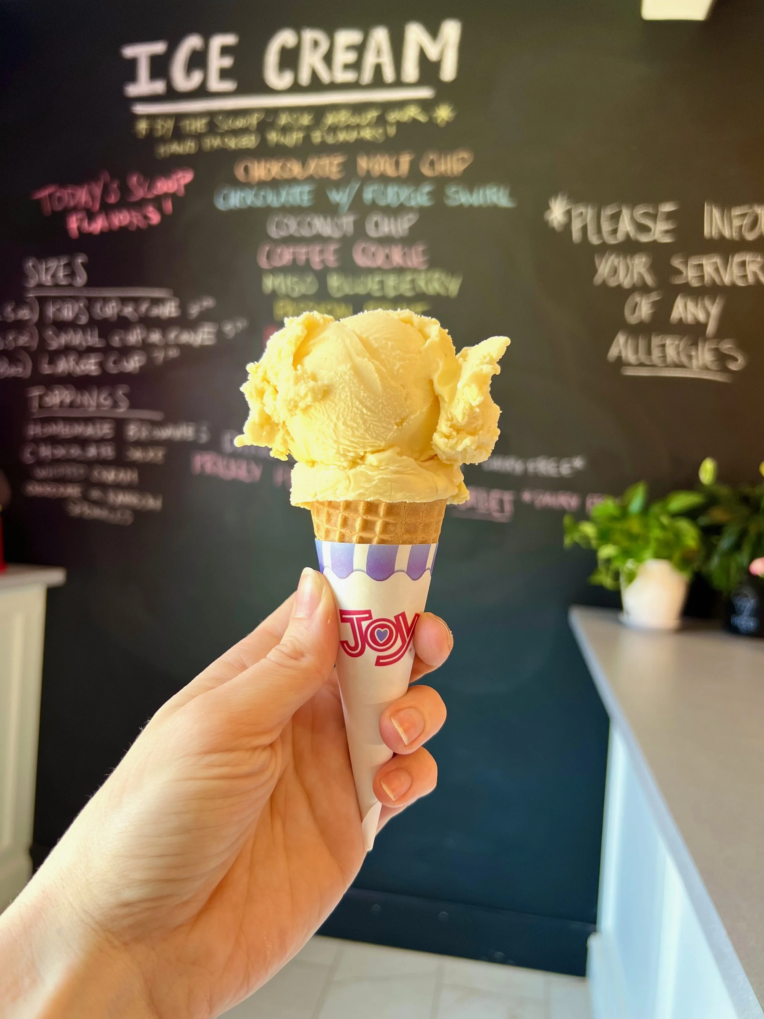 A person holds a scoop of homemade passionfruit ice cream in a cone with a Joy brand wrapper. In the background, there is a black chalkboard with colorful handwritten menu items related to ice cream flavors and toppings.