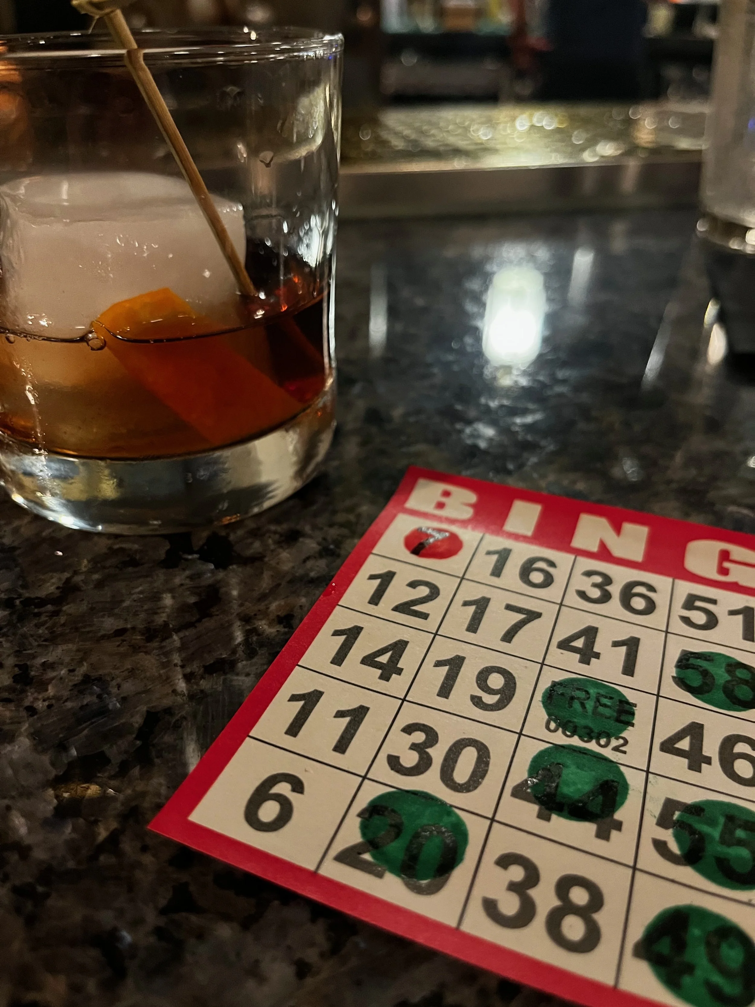 An old fashioned cocktail with a large ice cube and orange peel next to a red bingo card with green dauber marks on a granite bar top.