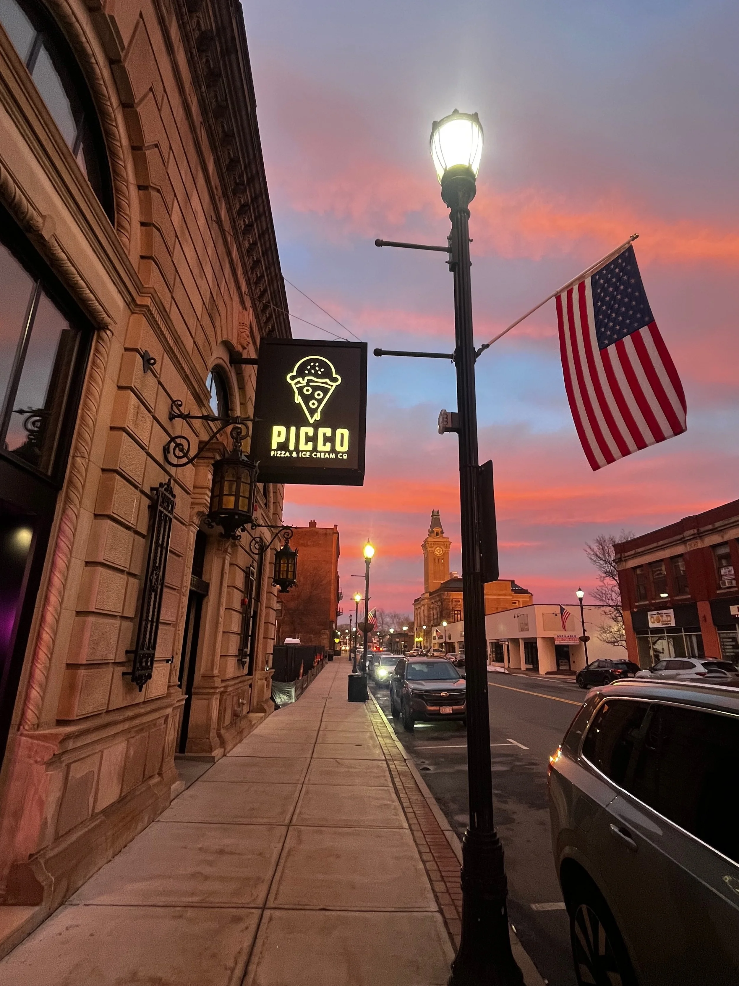 Street scene at dusk with a sidewalk, parked cars, historic building with ornate architecture on the left, a streetlamp illuminated, an American flag flying, and a sign for Pico Pizza & Ice Cream Co. against a colorful sky with pink and blue clouds.