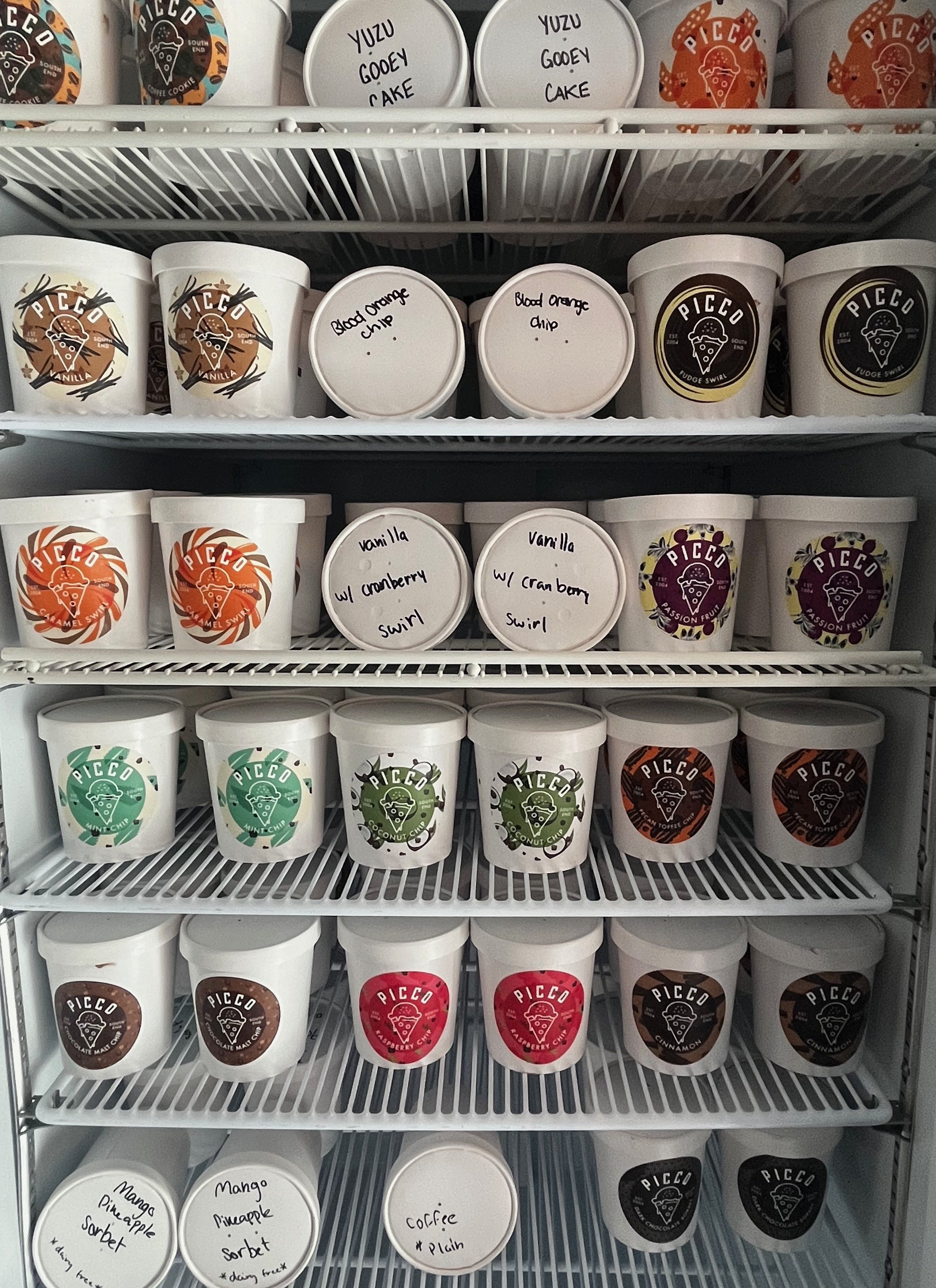 A refrigerated shelf containing various flavors of ice cream in Pintco brand containers. The flavors include Yuzu Gooey Cake, Vanilla with Cranberry Swirl, Passion Fruit, Mango Pineapple Sorbet, Coffee with Plum, and Cinnamon. Handwritten labels on the lids indicate the flavors.