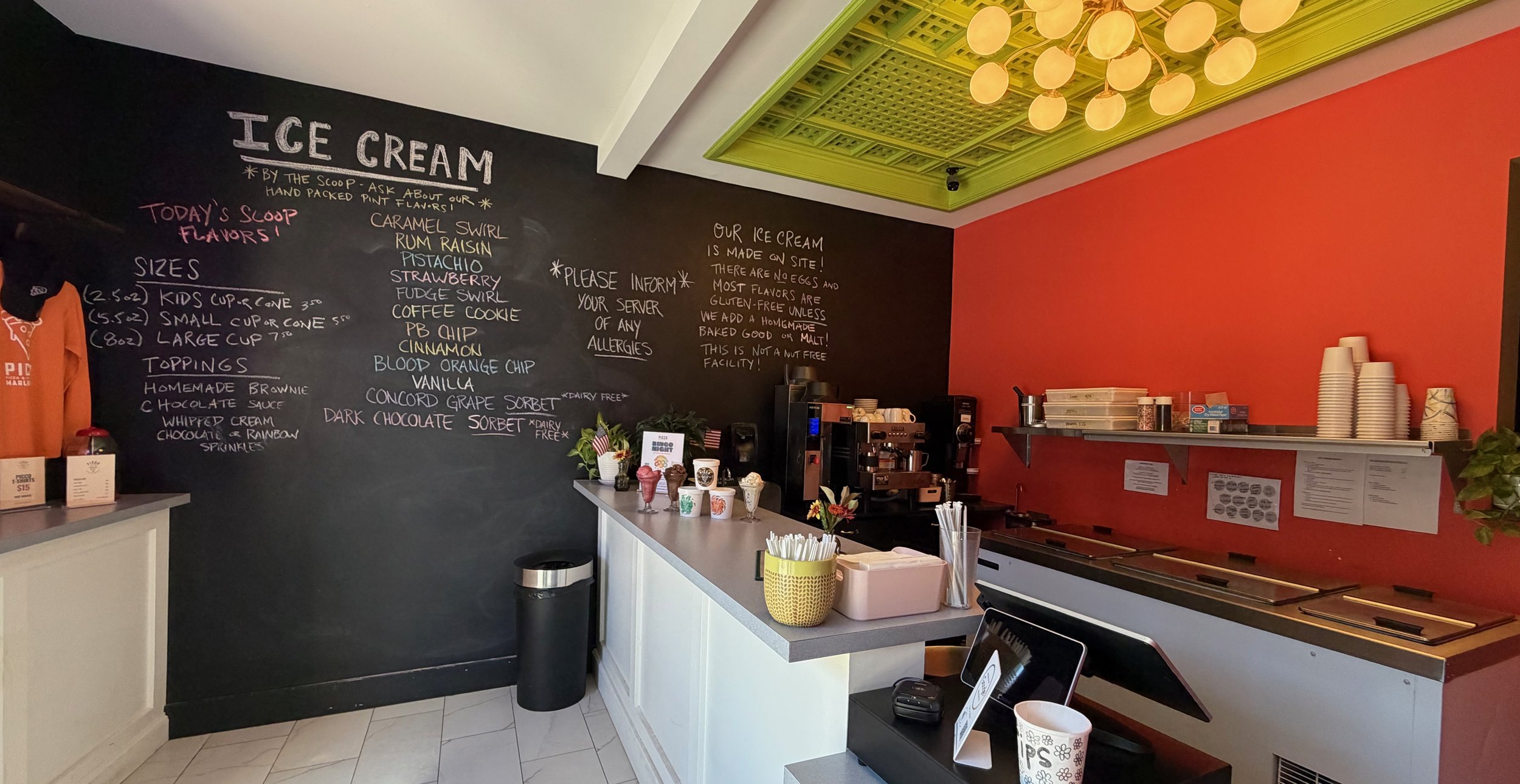 Counter at an ice cream shop with a black chalkboard menu on the wall listing flavors, sizes, and toppings; colorful ice cream cups on the counter, a coffee machine, and paper cups on shelves, with a decorative ceiling and a vibrant wall.