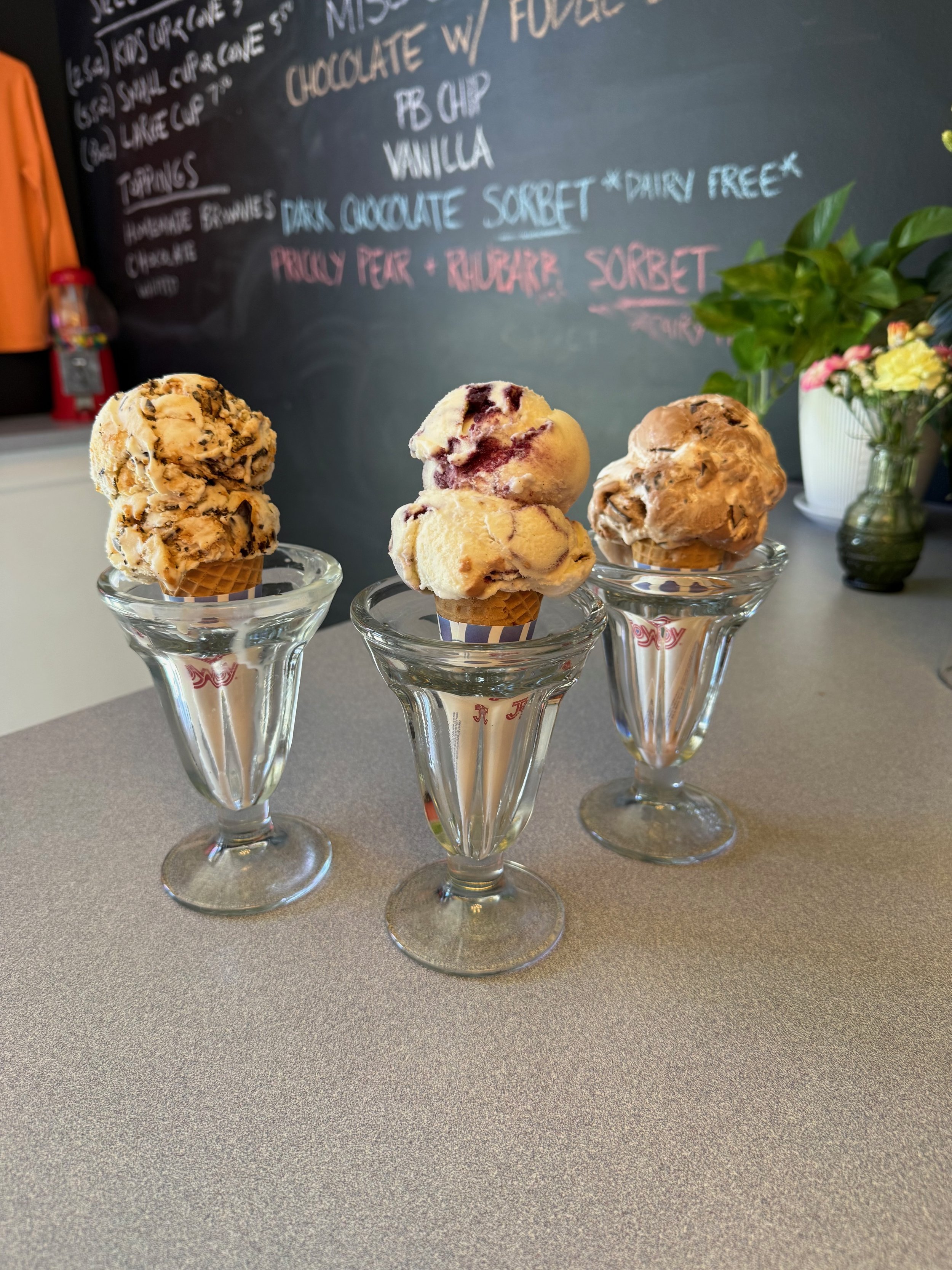 Three ice cream cones with different flavors in glass cups on a countertop, with a blackboard menu and a plant with flowers in the background.