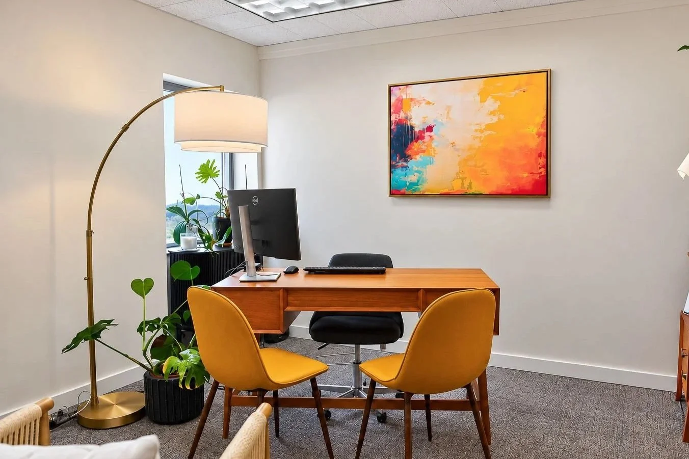 An office suite with a wooden desk, black office chair, potted plant, table lamp, and window with blinds.