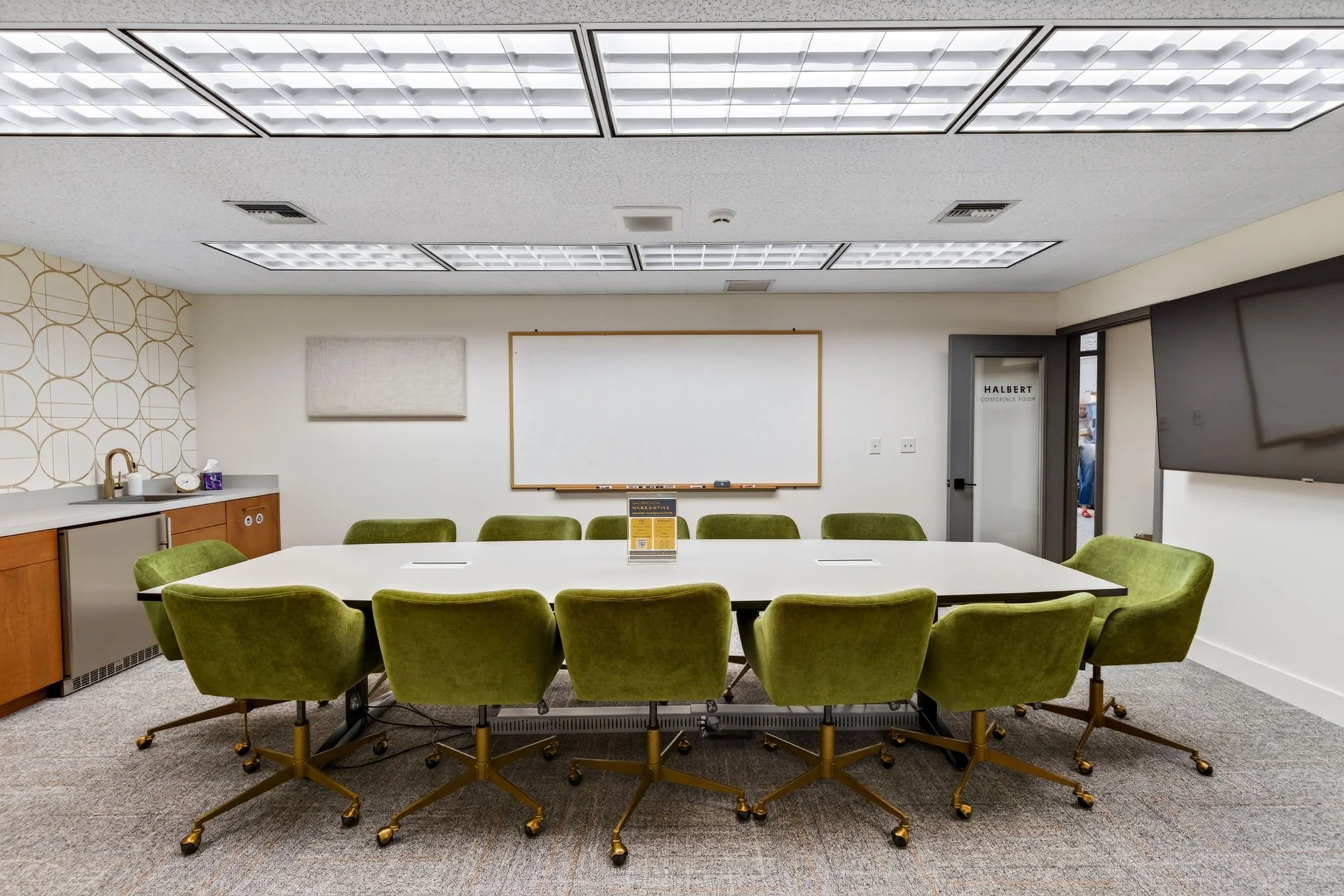 Conference room with a long white table surrounded by ten green upholstered chairs, a whiteboard on the wall, and a kitchenette with cabinets, a sink, a mini refrigerator, and a patterned wallpaper on one wall.