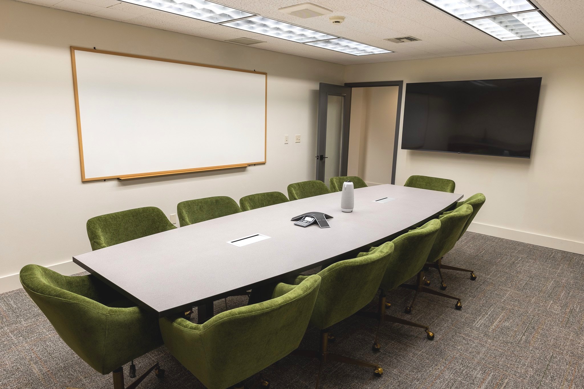 Conference room with a large rectangular table surrounded by green office chairs, a whiteboard on the wall, a large flat-screen TV, and conference call equipment in the center of the table.