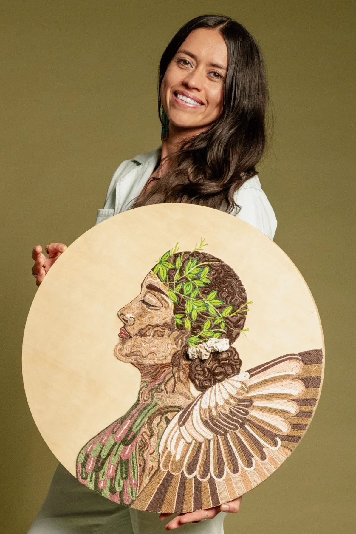 A woman with long dark hair and a white shirt smiling, holding a round artwork depicting a woman with leaves and flowers in her hair and butterfly wings.