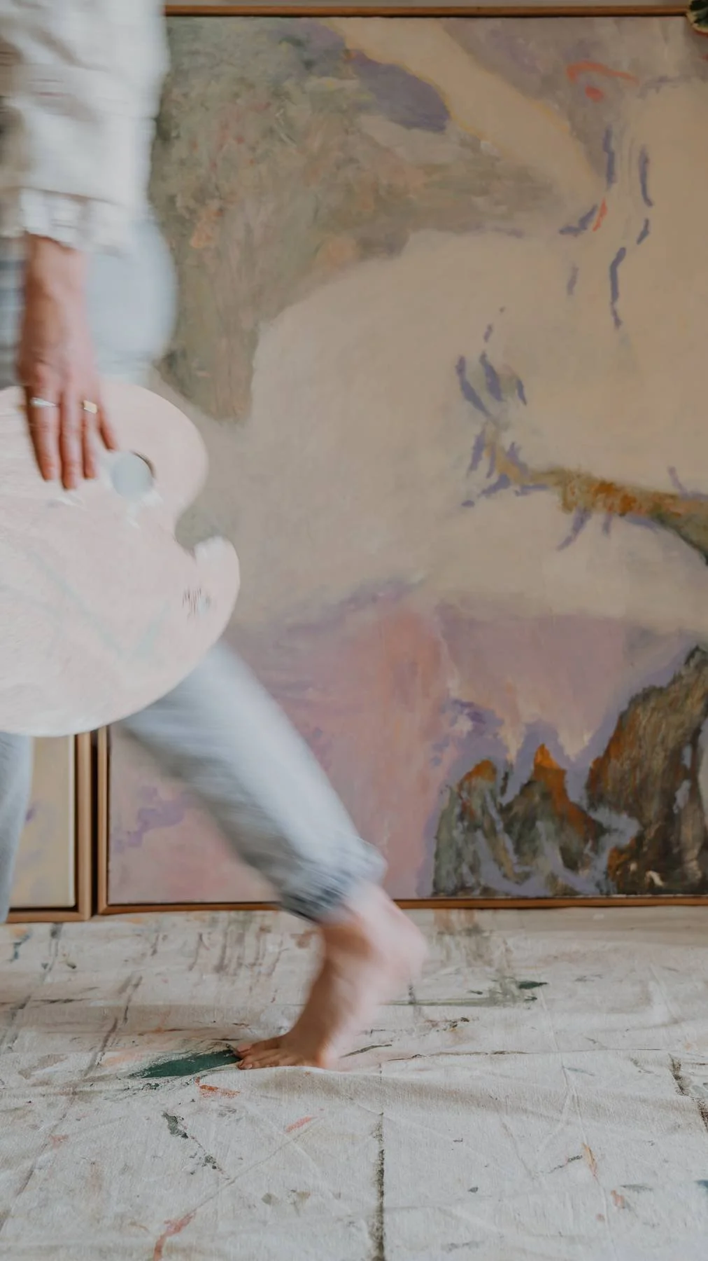 Person in a white shirt and patterned pants capturing an abstract painting with one hand, while balancing on one foot on a cloth-covered floor.
