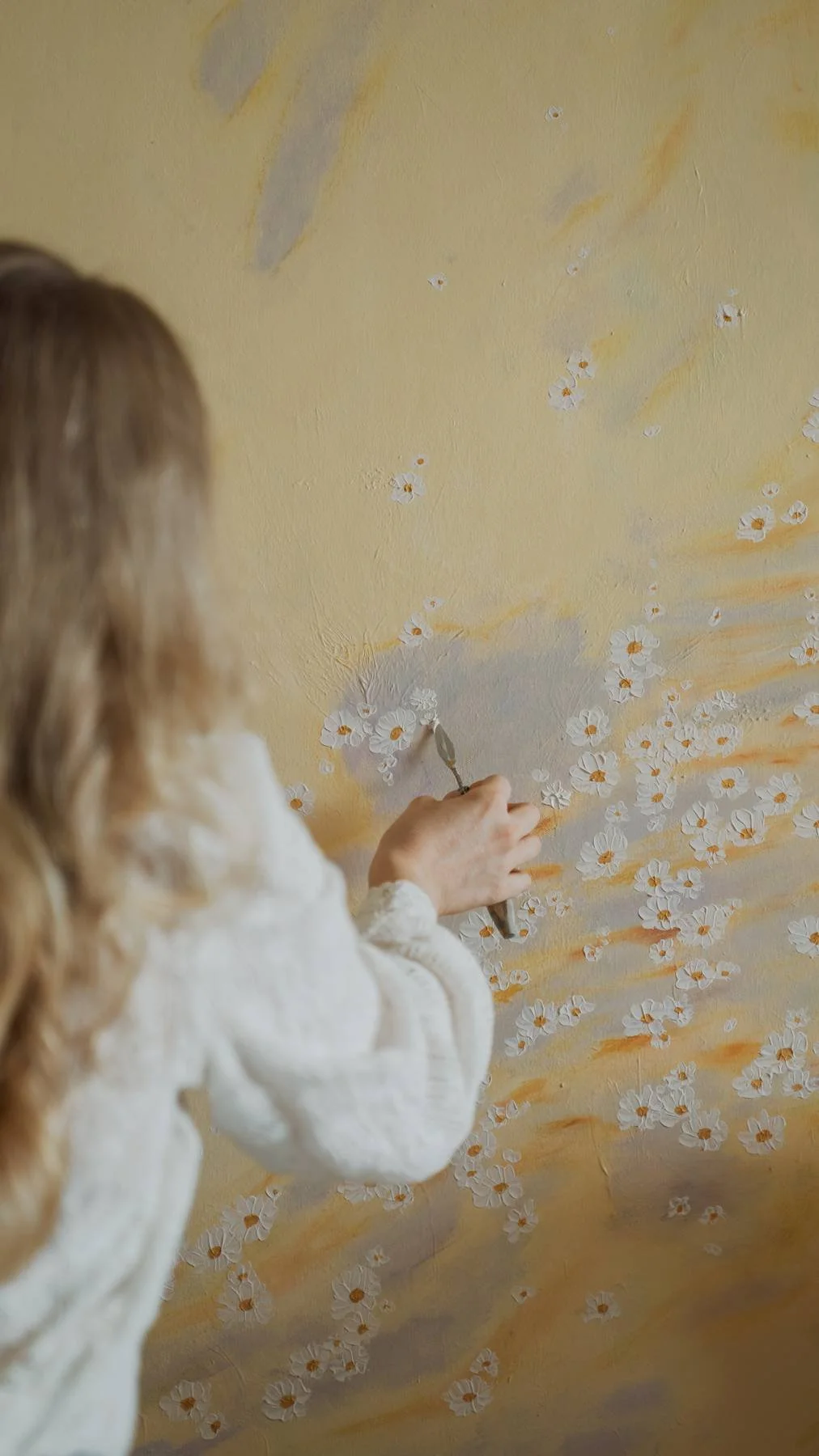 A person with long curly hair in a white sweater is painting white and orange flowers on a yellow wall.