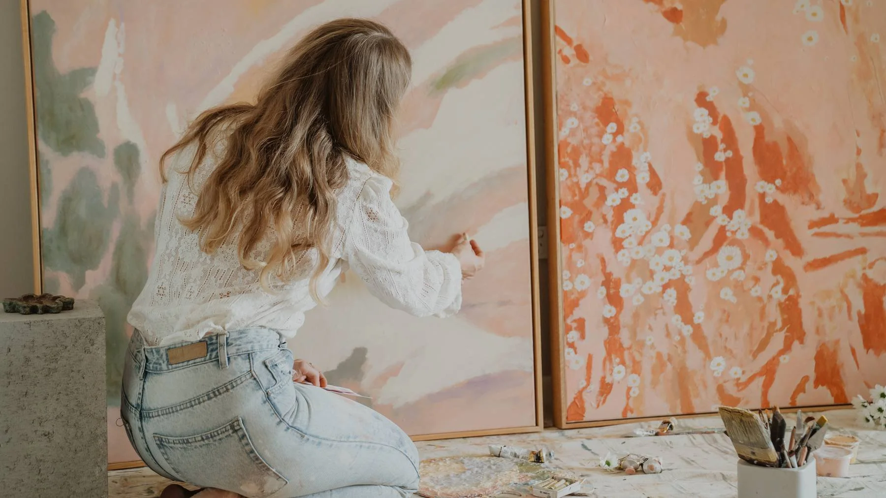An artist with long hair painting in a studio, surrounded by large abstract paintings and art supplies on the floor.