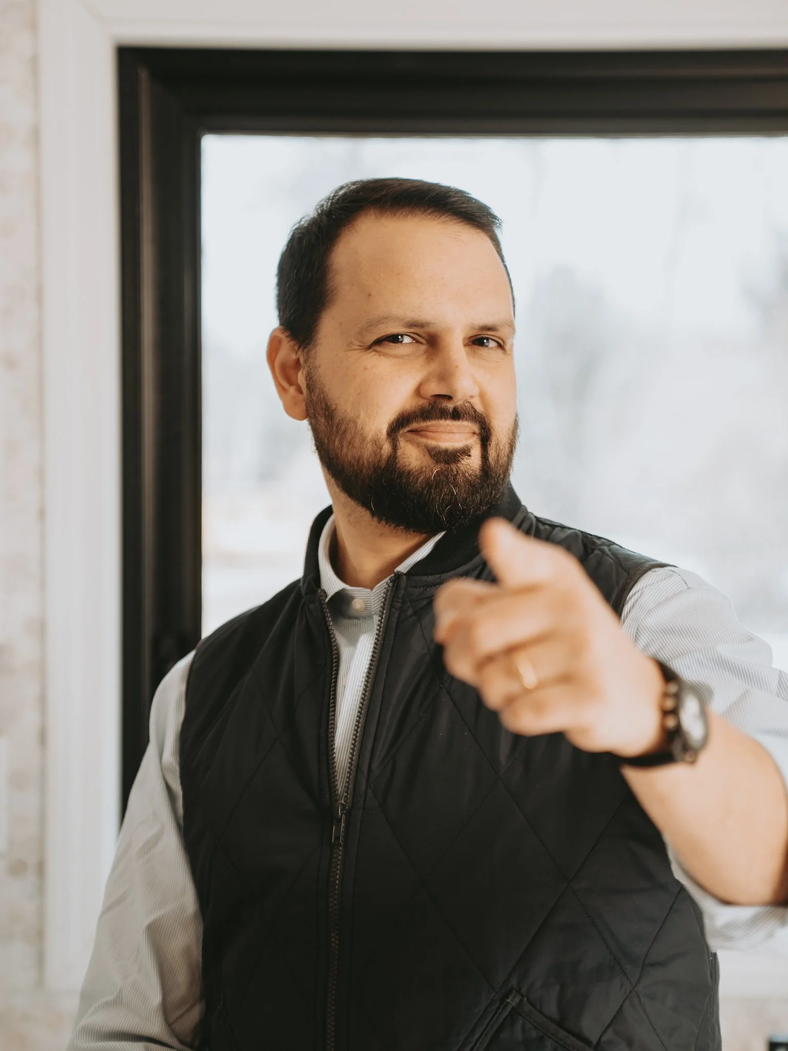 A confident man with a beard and short dark hair pointing towards the camera inside a house near a window.