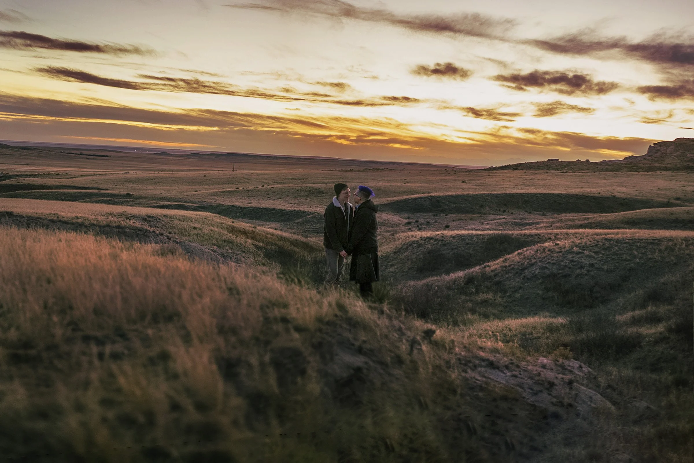 Two people standing together on a grassy hill during sunset, with a vast landscape in the background.