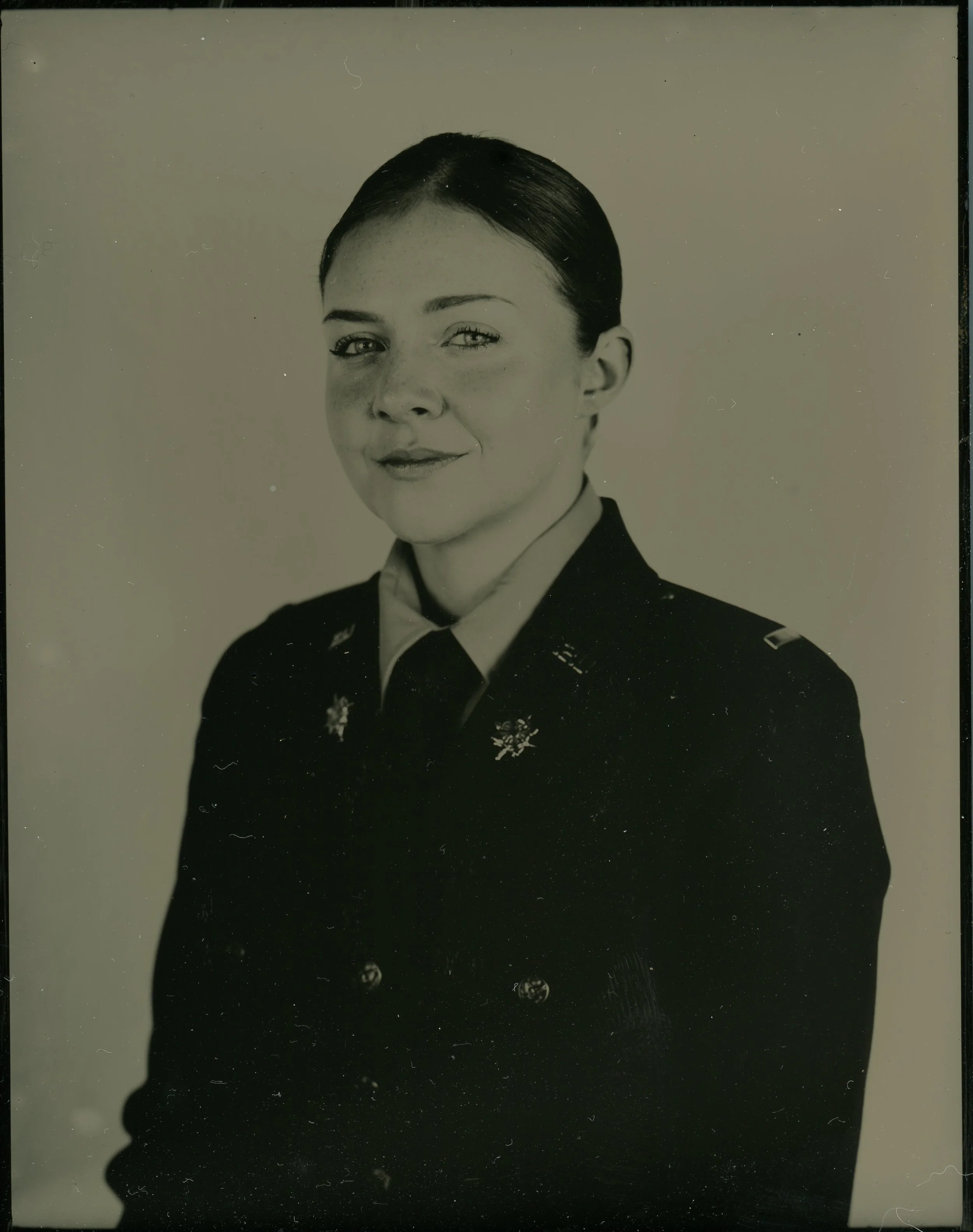 A black and white portrait of a person in a military uniform with insignia, looking at the camera. The uniform has shoulder epaulettes and decorative pins.