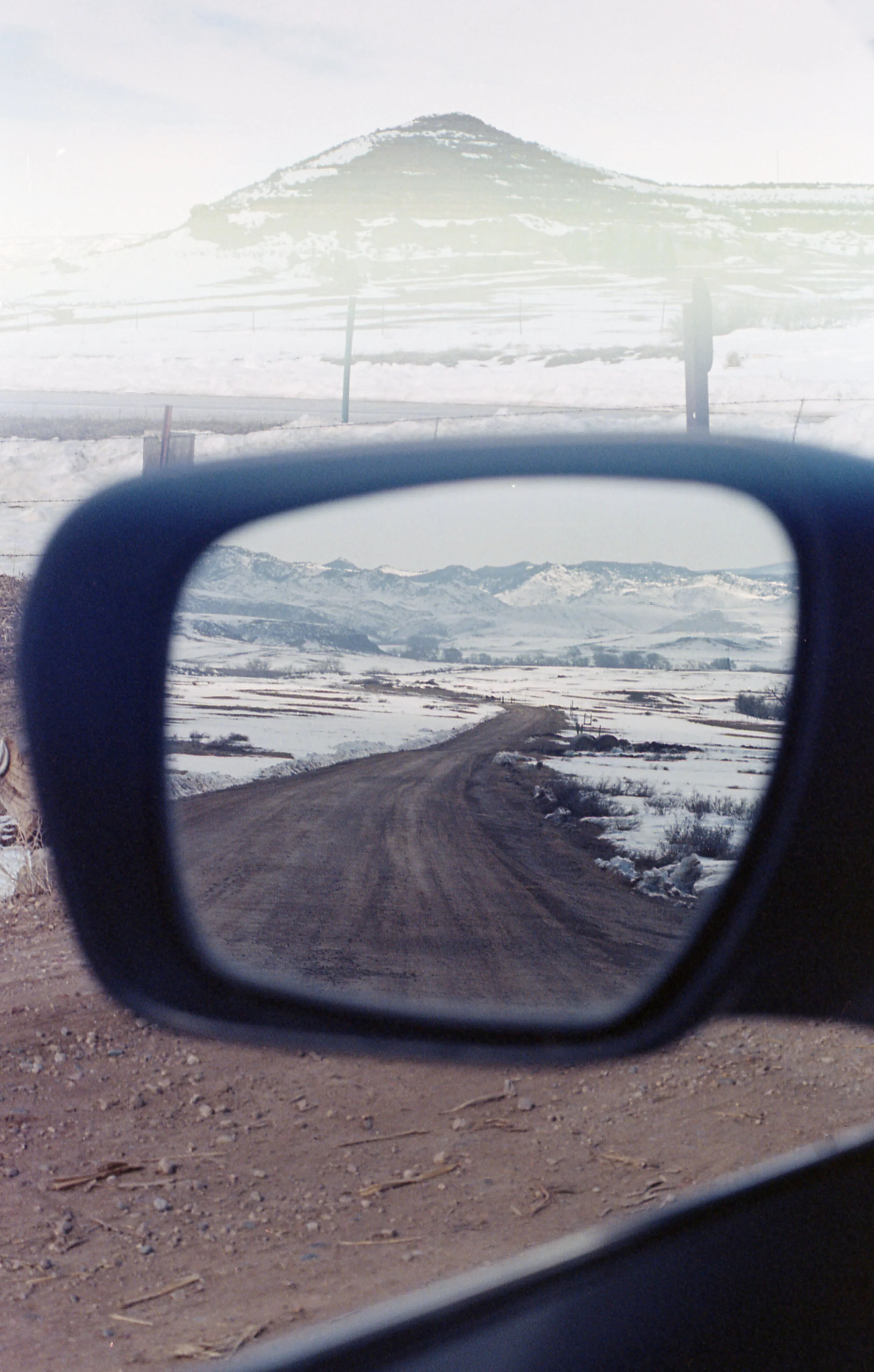 Snowy landscape reflected in car side mirror with a dirt road and mountain in view.