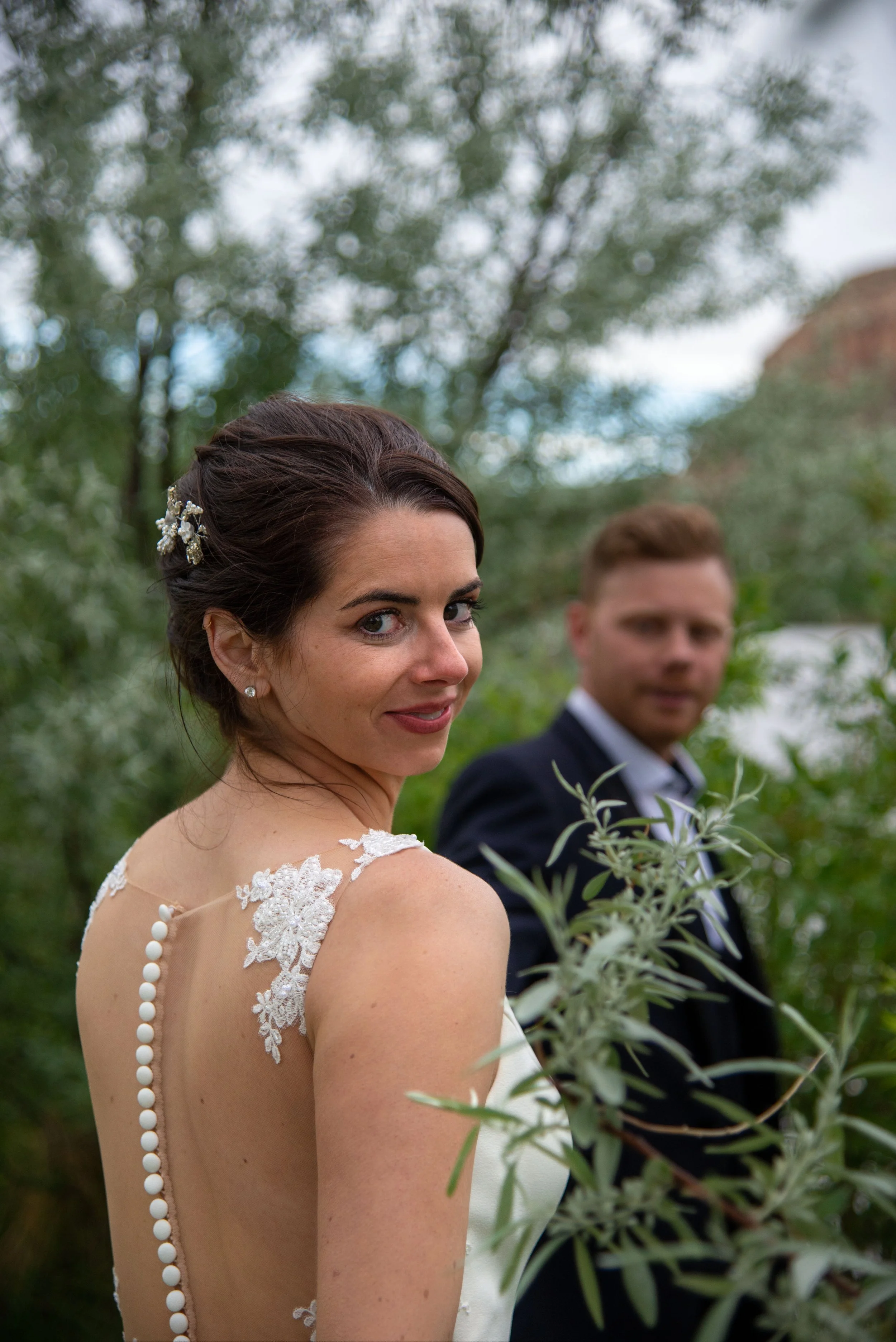 Bride in lace wedding dress smiling with groom in background, surrounded by greenery.