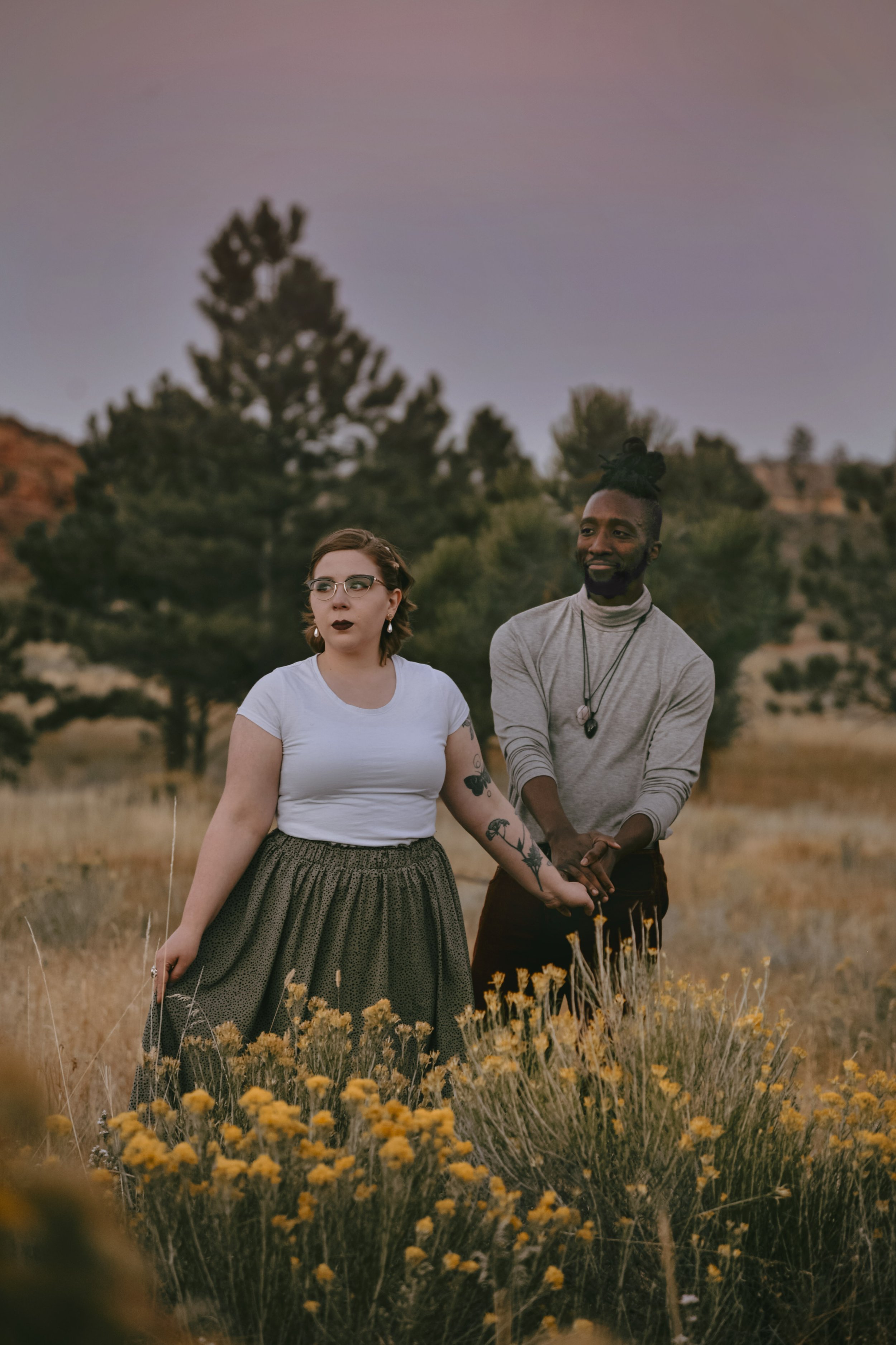A man and woman holding hands in a field with yellow flowers and trees, under a dusky sky.