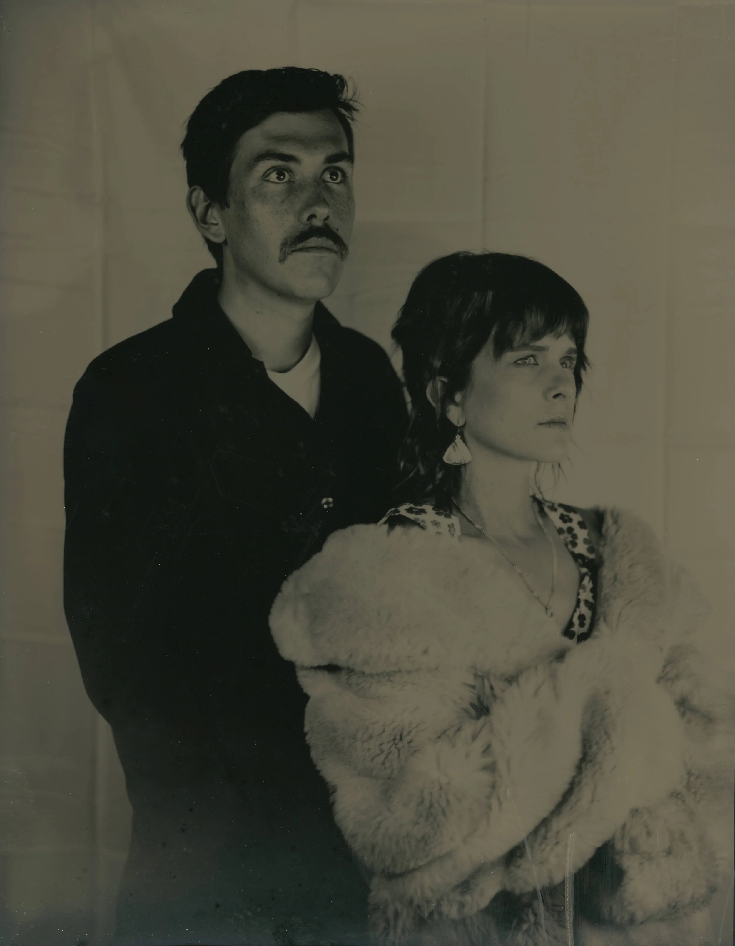 Black and white photo of a man with a mustache and dark hair, and a woman with shoulder-length dark hair, wearing earrings and a fur coat, standing in front of a tiled wall.