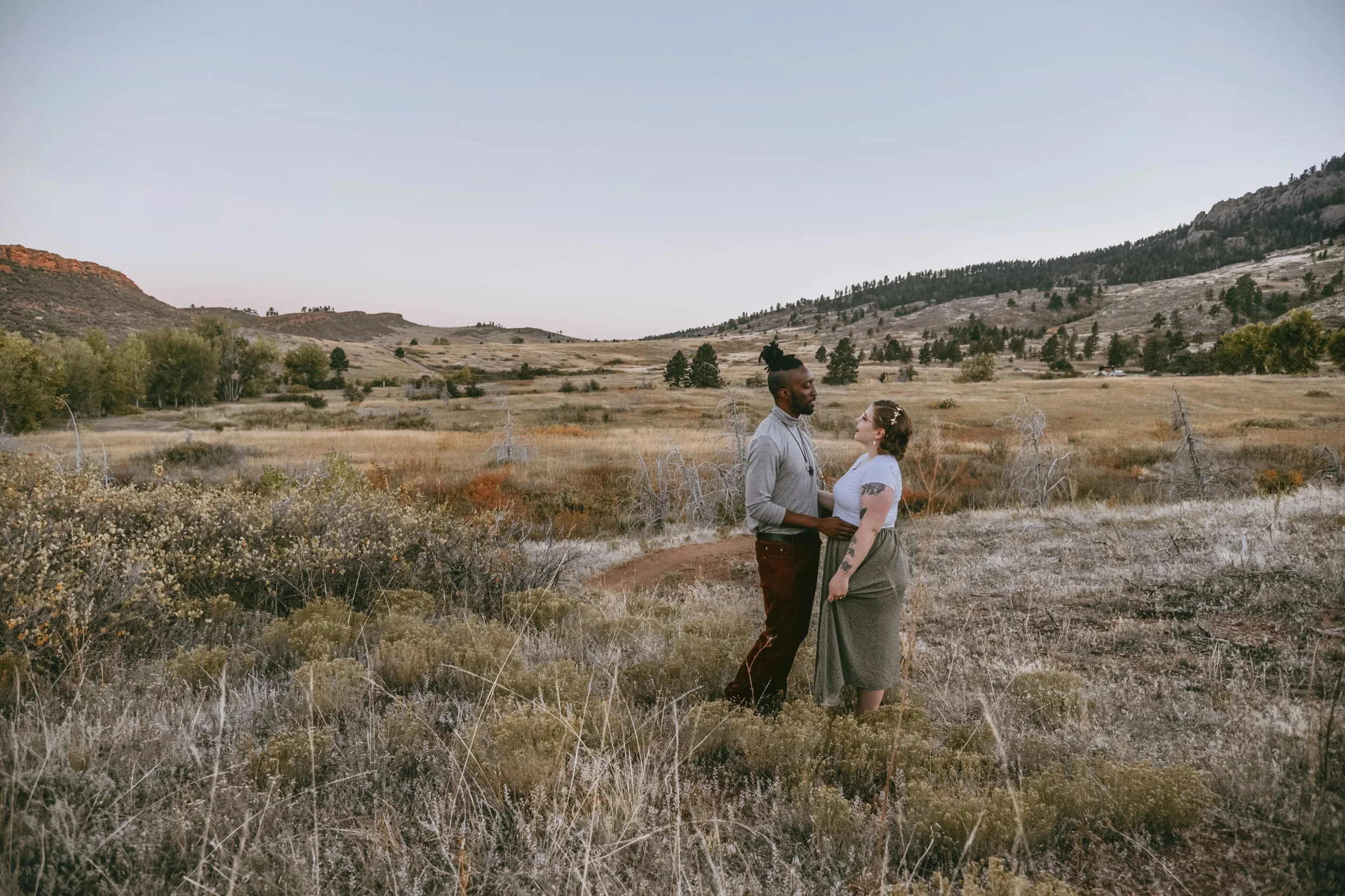 Couple standing in a scenic meadow with hills and scattered trees in the background, under a clear sky.