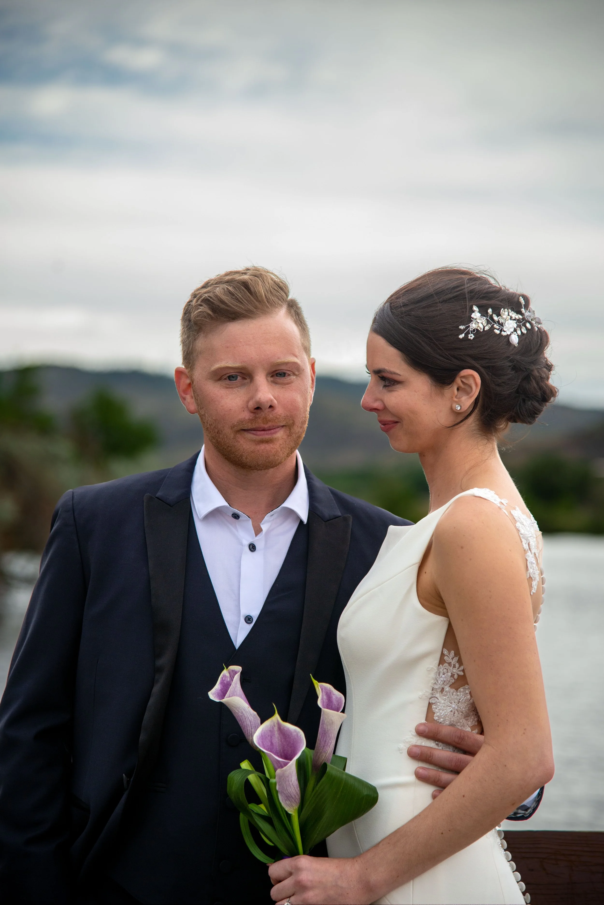 Bride and groom standing together outdoors, bride holding calla lilies, wearing white dress, groom in black suit with vest, outdoor backdrop of trees and sky.