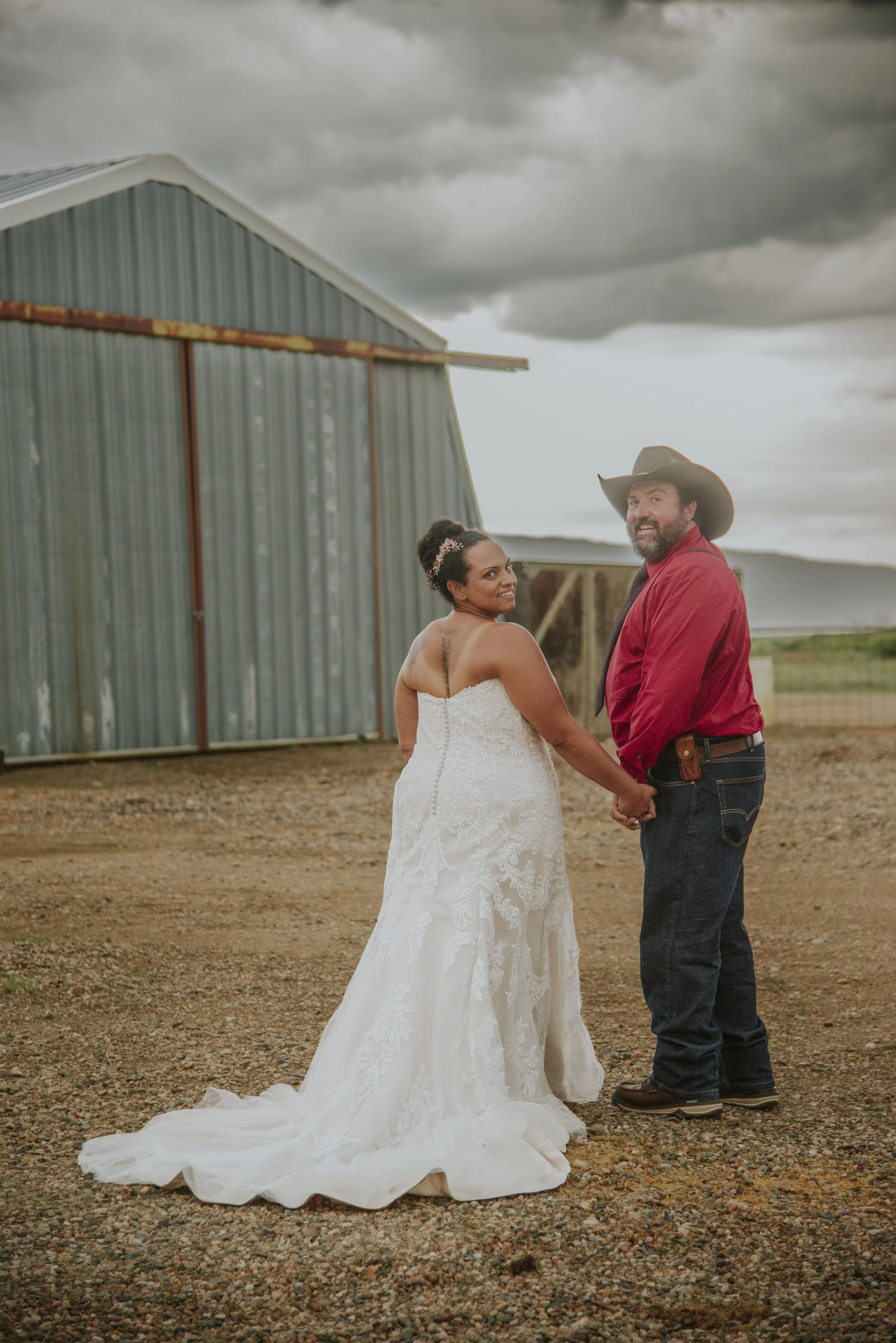 Bride in a white dress and groom in a red shirt with a cowboy hat, holding hands in front of a metal barn, outdoor setting, cloudy sky.