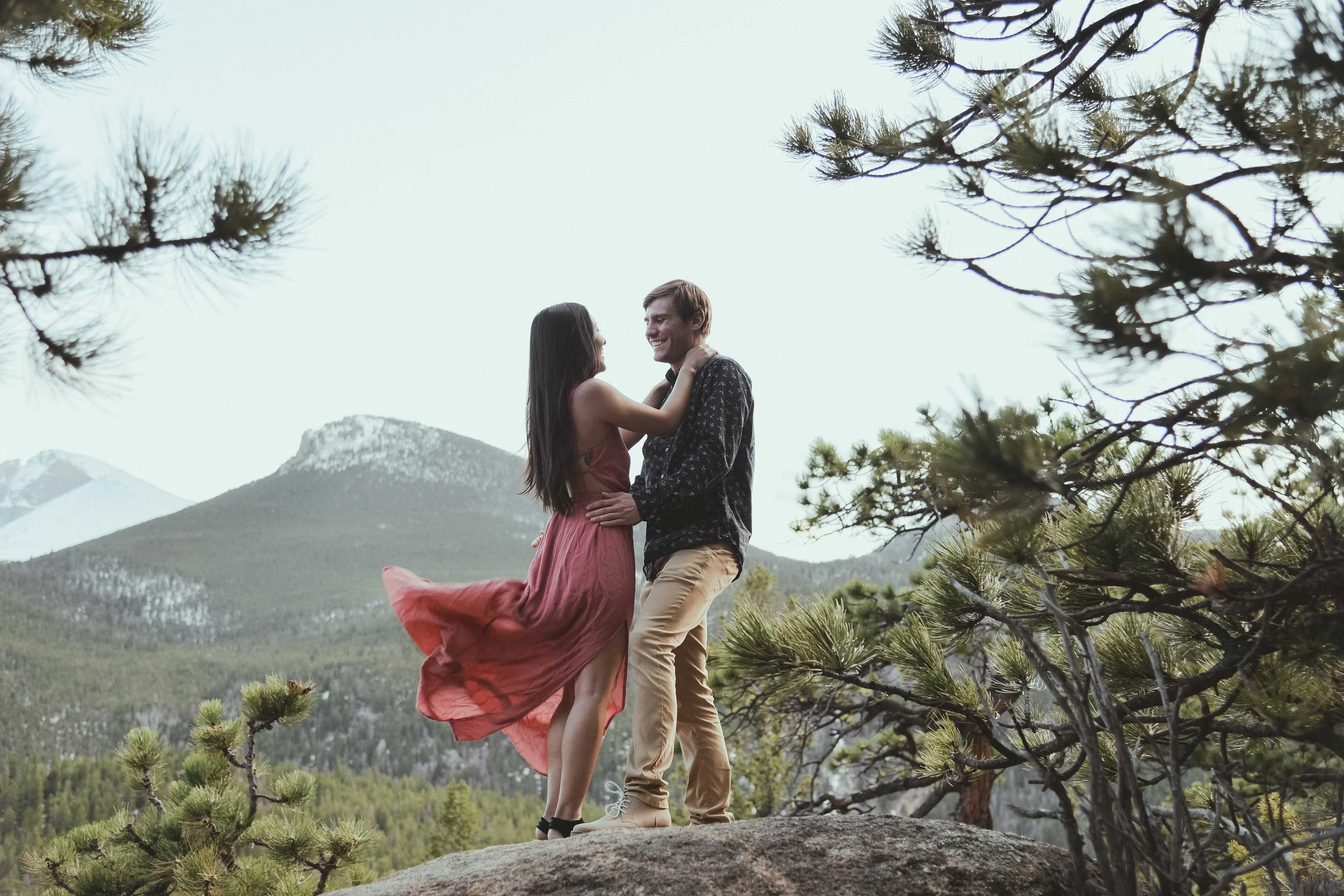 Couple embracing on a rocky hilltop with mountainous landscape and trees.