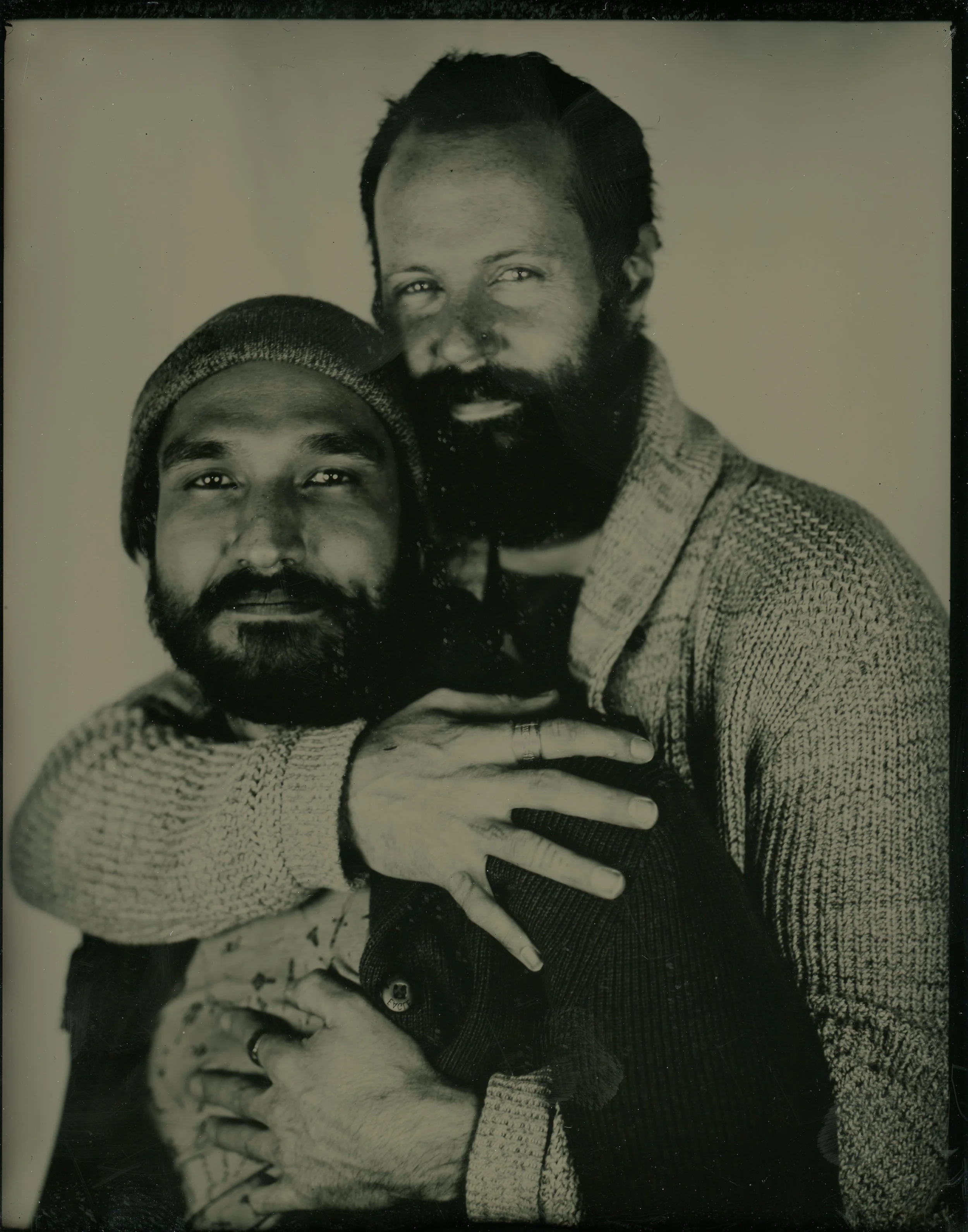 Two men with beards embrace, one wearing a beanie and the other a knit cardigan, in a vintage-style sepia photograph.