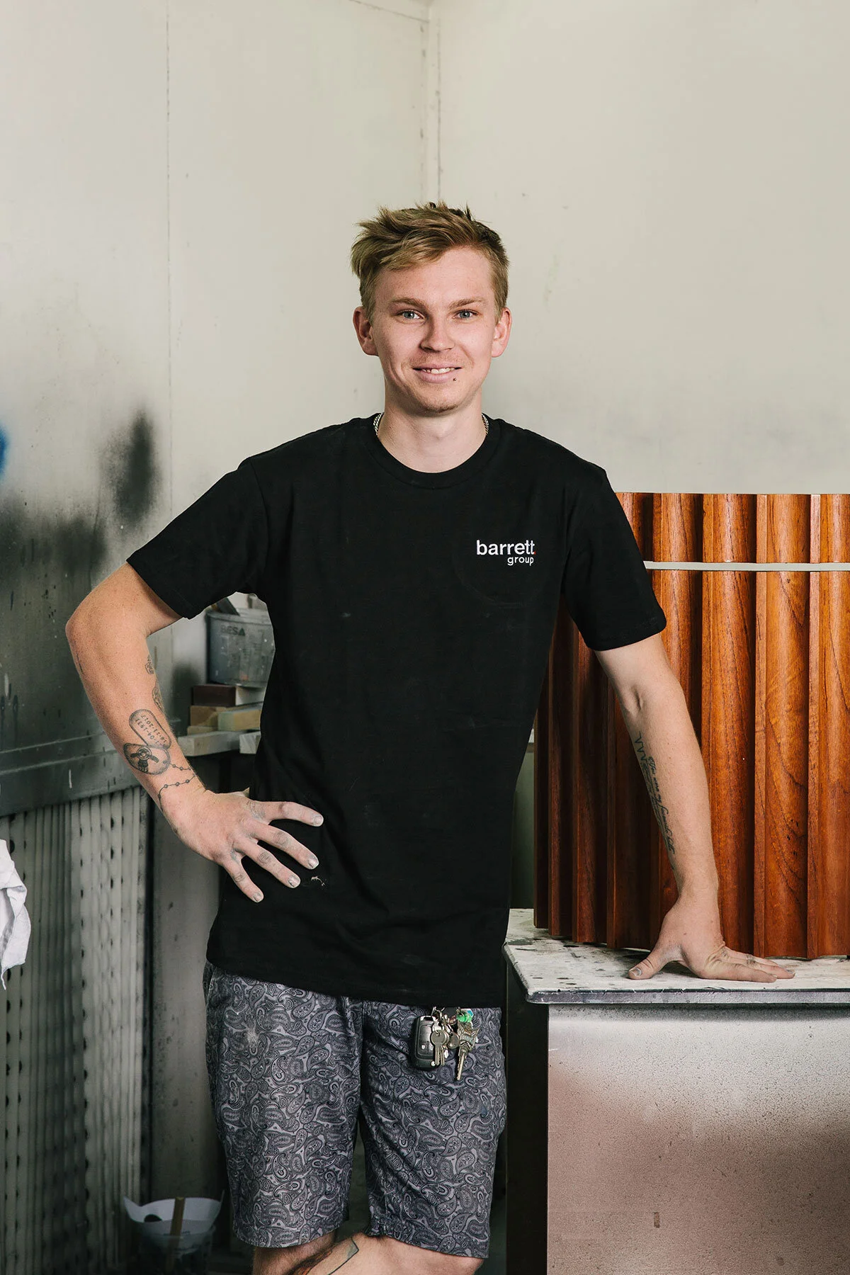 A young man with blonde hair, wearing a black t-shirt with 'barrett group' logo, stands with one hand on his hip and the other resting on a wooden structure. He is smiling in a casual setting, with tattoos on his arms and keys hanging from his shorts.