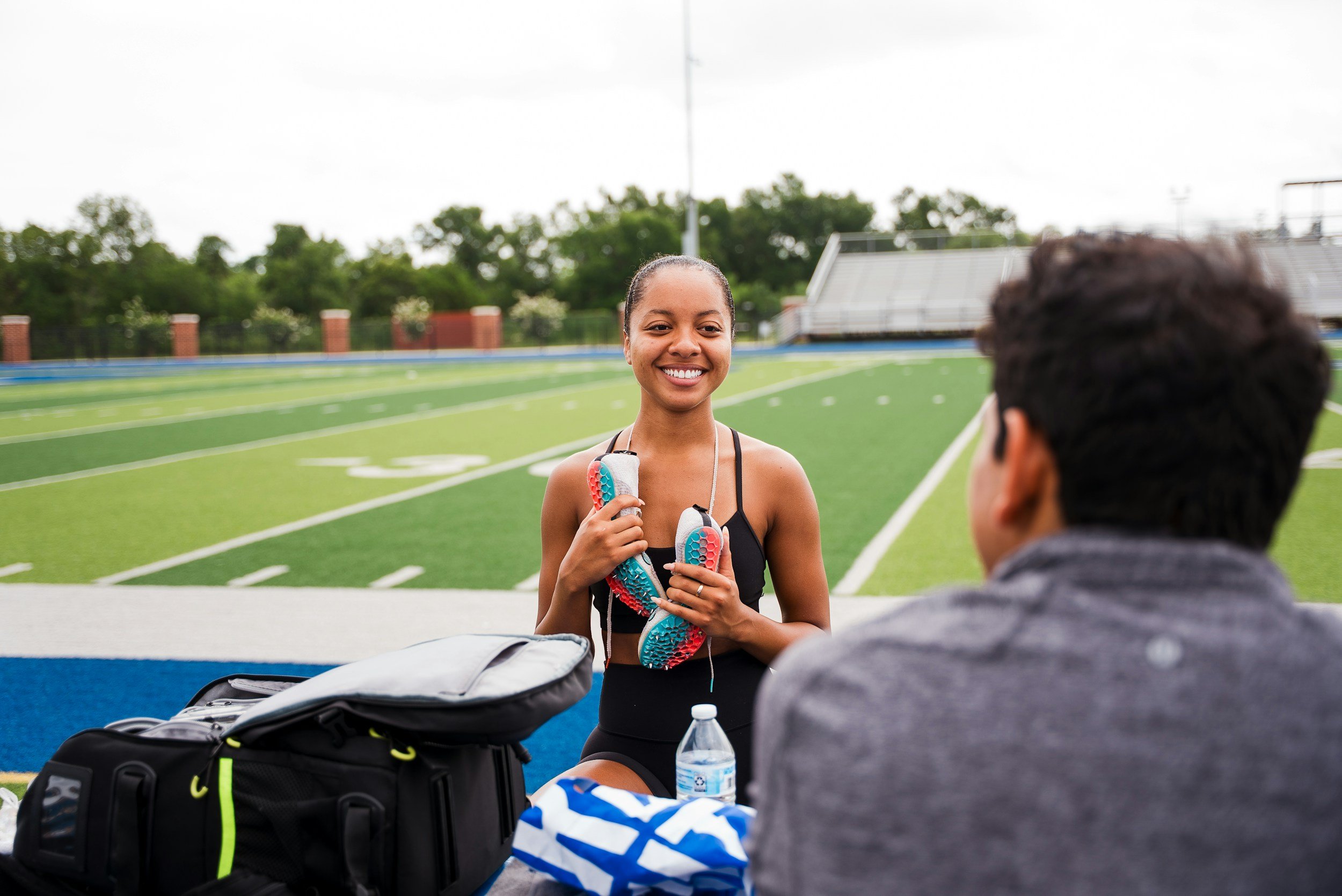 A smiling young woman holds new athletic shoes while sitting at an outdoor track and field stadium, with a man seated across from her. There are backpacks and water bottles on the table in front of them.