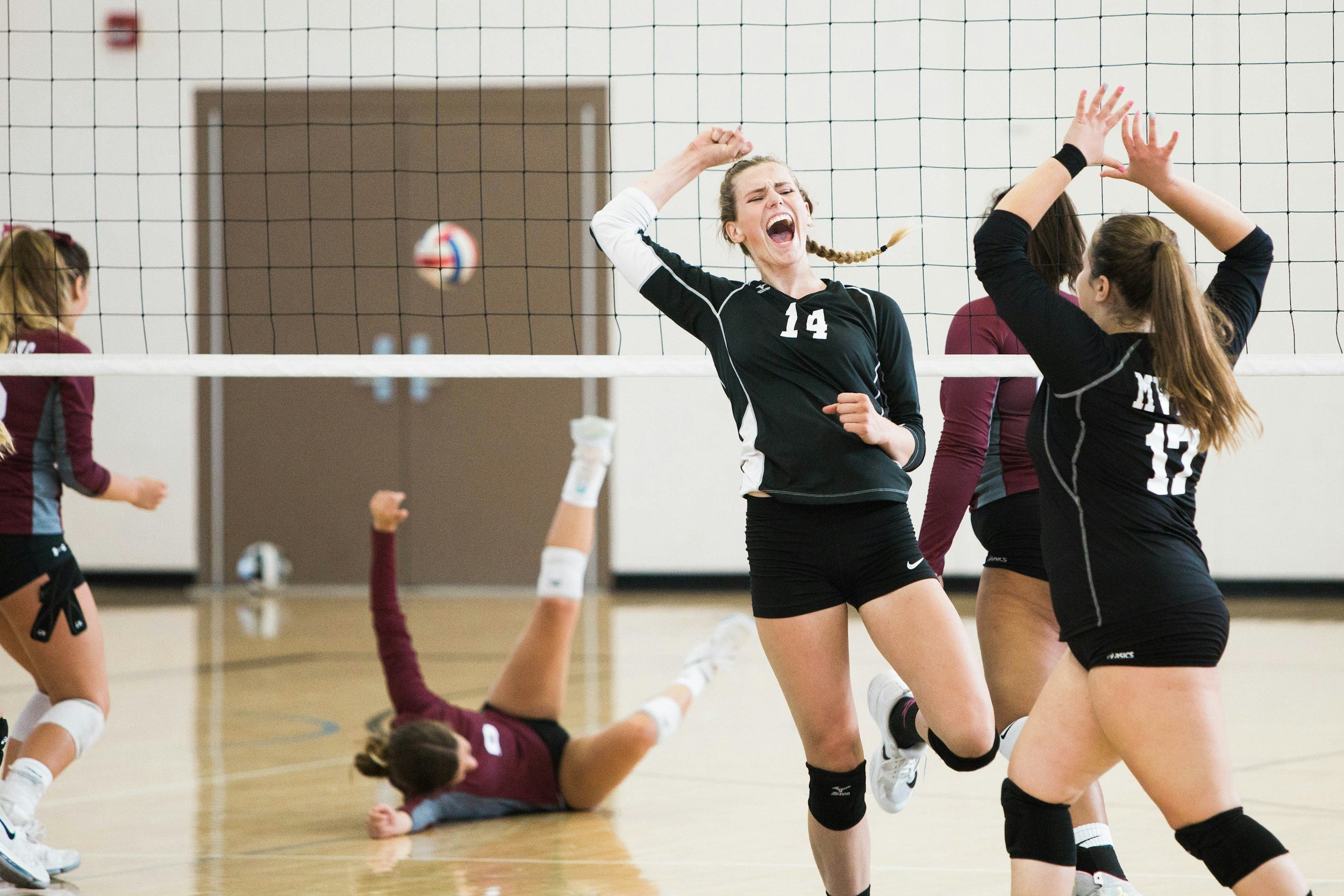 Volleyball players celebrating on the court after a point, with a player lying on the floor and others standing near the net.