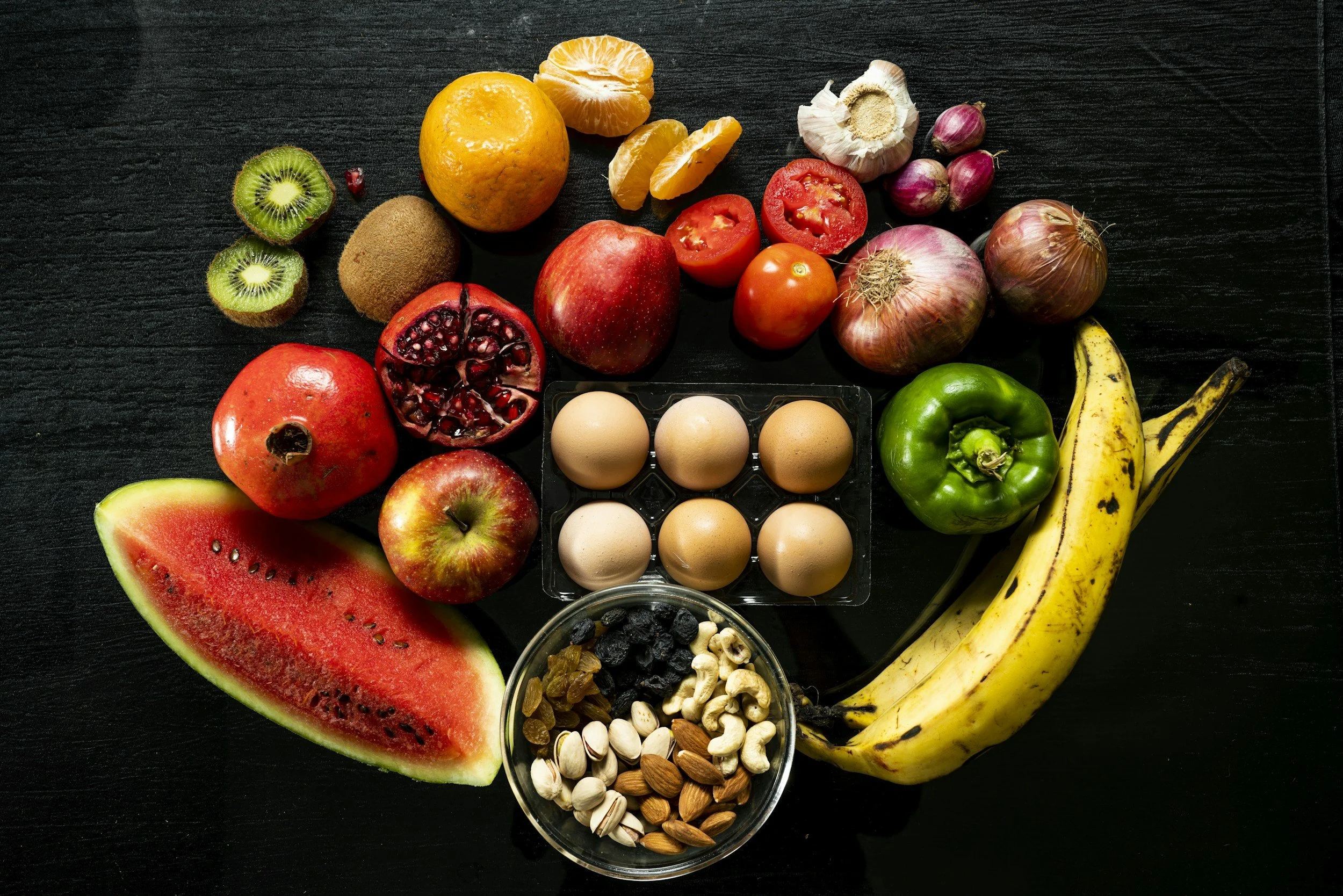 assortment of fresh fruits and eggs on a black surface, including watermelon, kiwi, tangerines, oranges, pomegranate, apples, tomatoes, garlic, onions, bananas, and a bowl of mixed nuts and dried fruits.