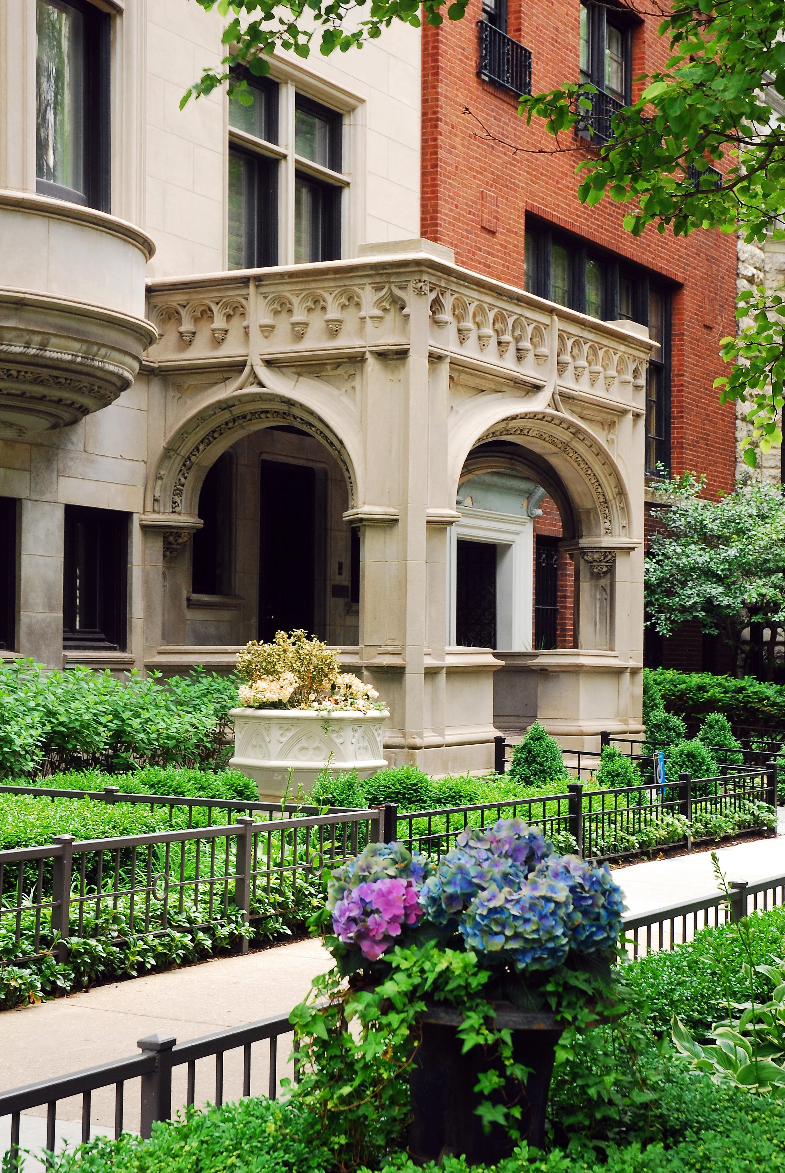 Ornate stone entrance of a building with arches and decorative details, surrounded by neatly manicured greenery and flowering plants, including hydrangeas, in an urban garden setting.