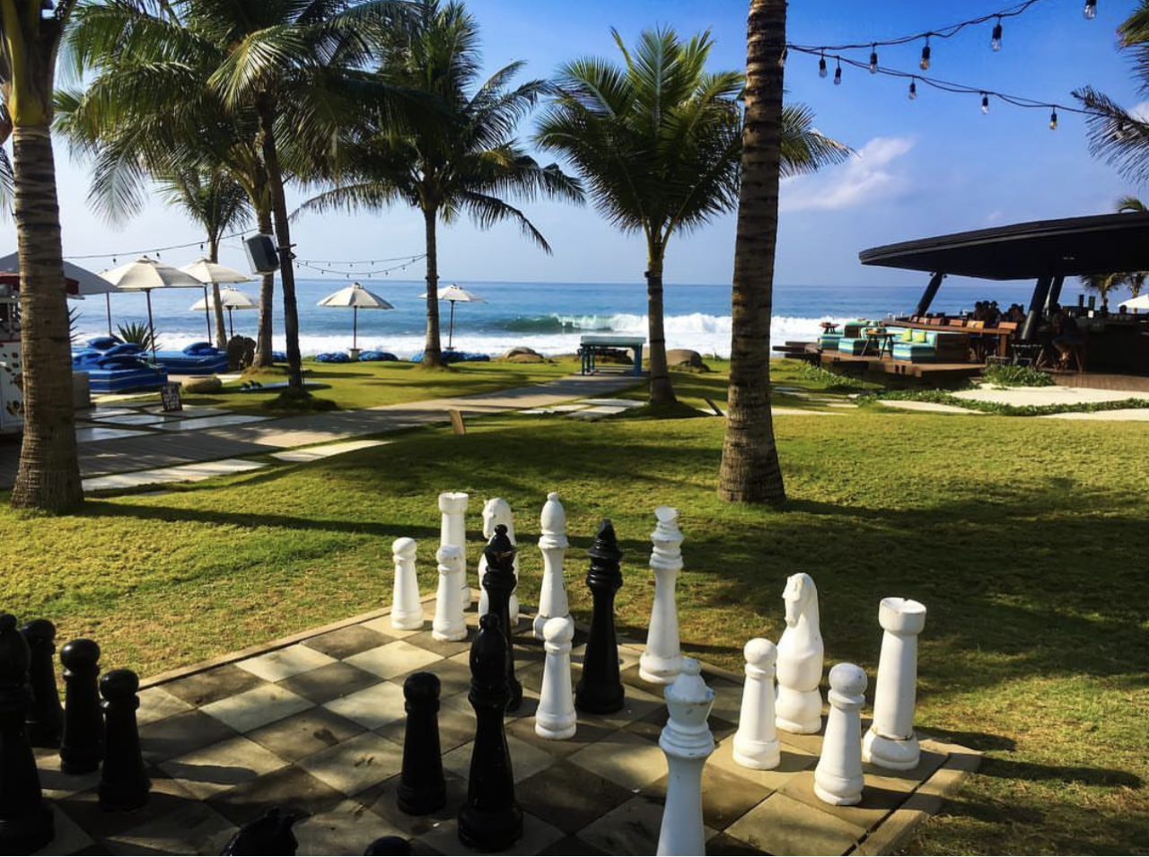 A scenic view of a beachside resort with chess pieces on a chessboard in the foreground, palm trees, umbrellas, lounge chairs, a bar, and the ocean in the background.