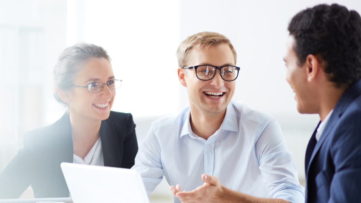 Three people having a conversation in a bright office, two women and one man, smiling and engaged in discussion.