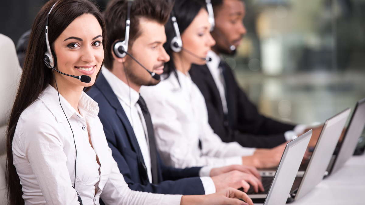 Four customer service representatives wearing headsets working at laptops in an office.