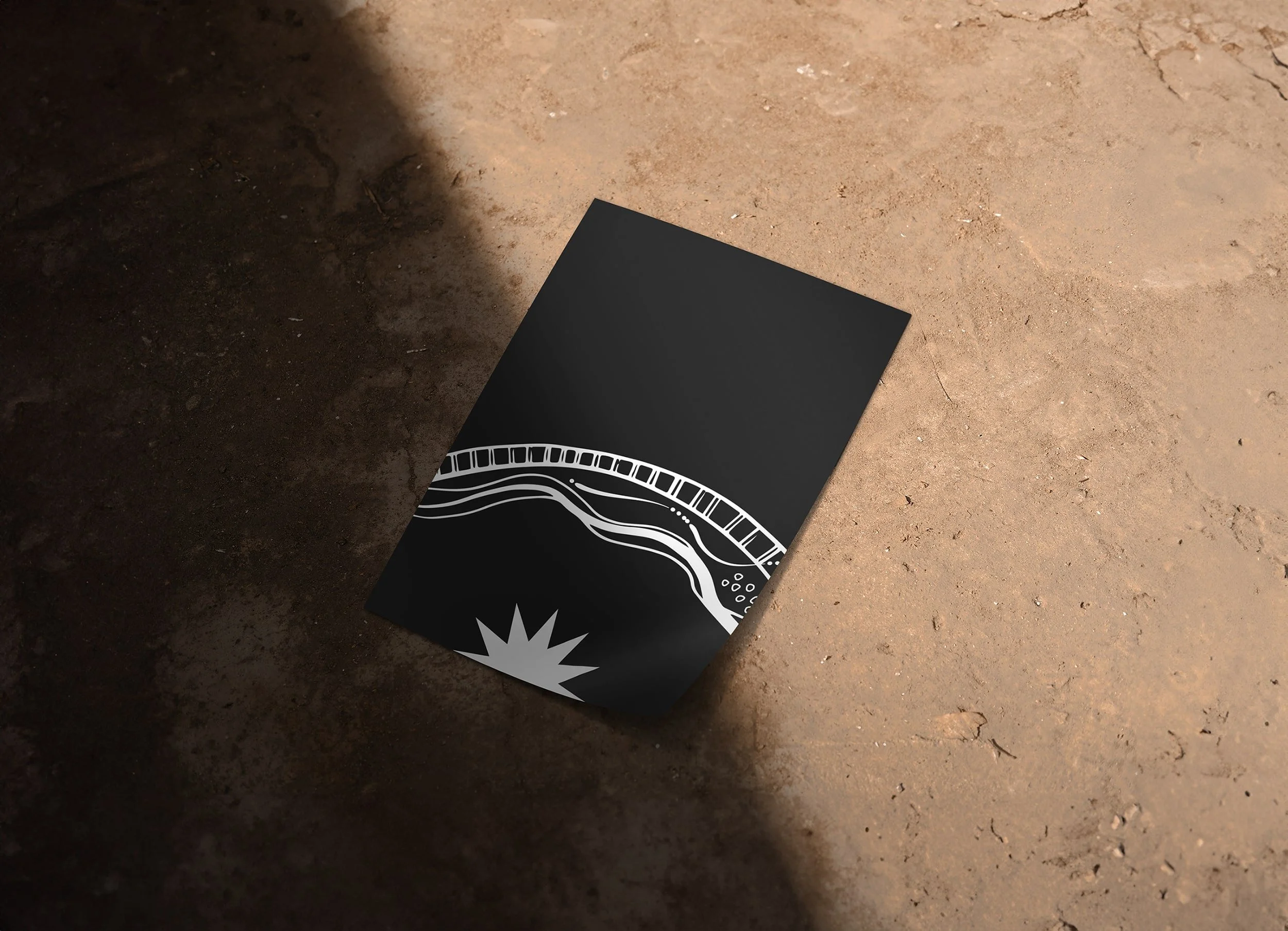 A black piece of paper with white abstract designs lying on a dirt surface, partially in shadow.