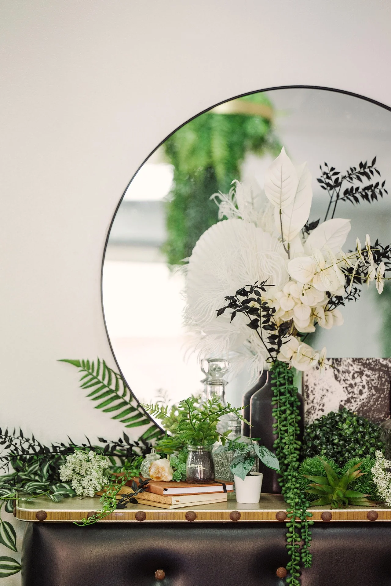 Decorative scene with a large round mirror, a variety of green and white artificial plants, a black vase, a crystal-like object, and stacked books on a vintage table.