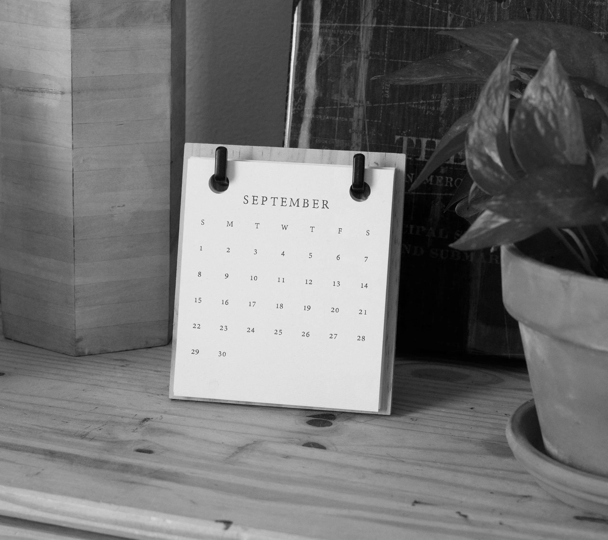 A black-and-white photo of a desk with a small calendar showing the month of September, plants, and a wooden box.