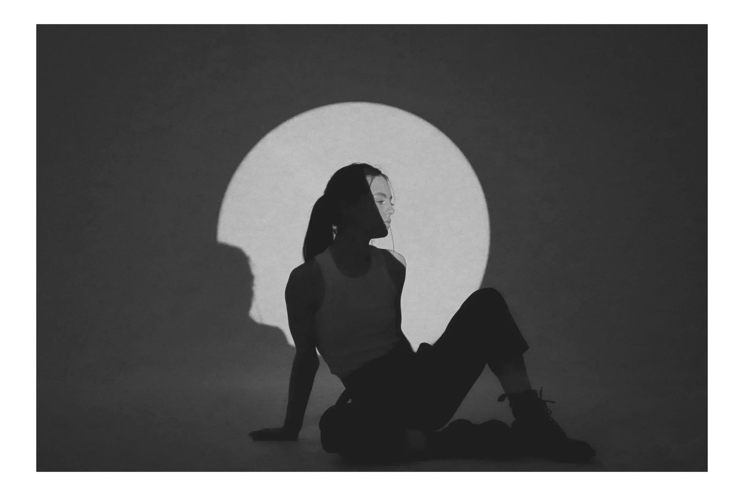 A black and white photo of a woman sitting on the floor, with her right arm supporting her and her left arm resting on her knee, against a backdrop of a large circular light. Her silhouette creates a shadow on the wall, and she gazes to the side with her hair tied back. The scene has a minimalist and artistic feel.