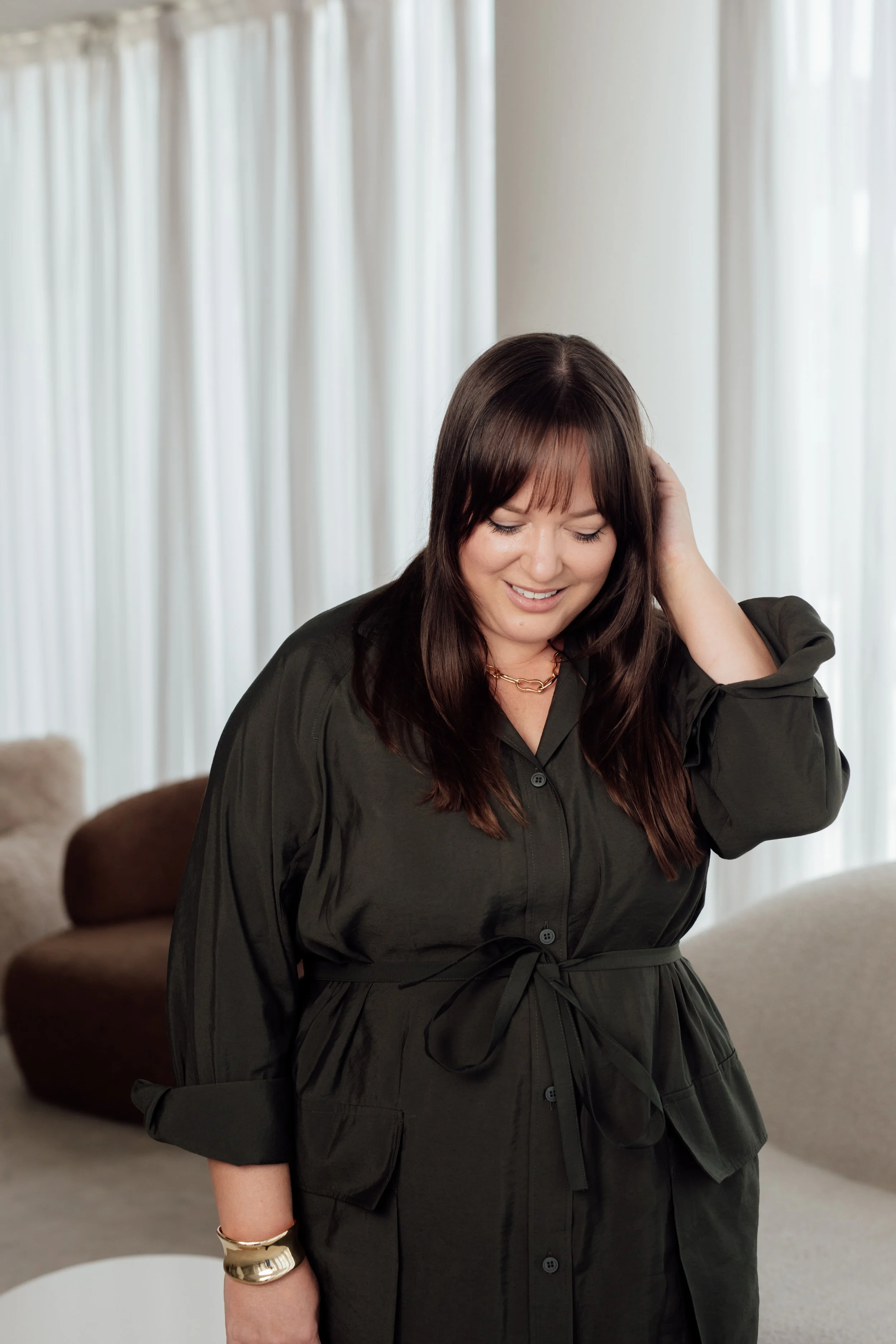 A woman with dark brown hair smiling, touching her hair, wearing a dark green dress with a waist tie, in a living room with light-colored curtains and a brown and a beige couch.