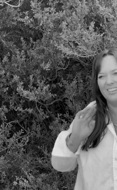 Laura Boatman standing outdoors near bushes, smiling and waving.