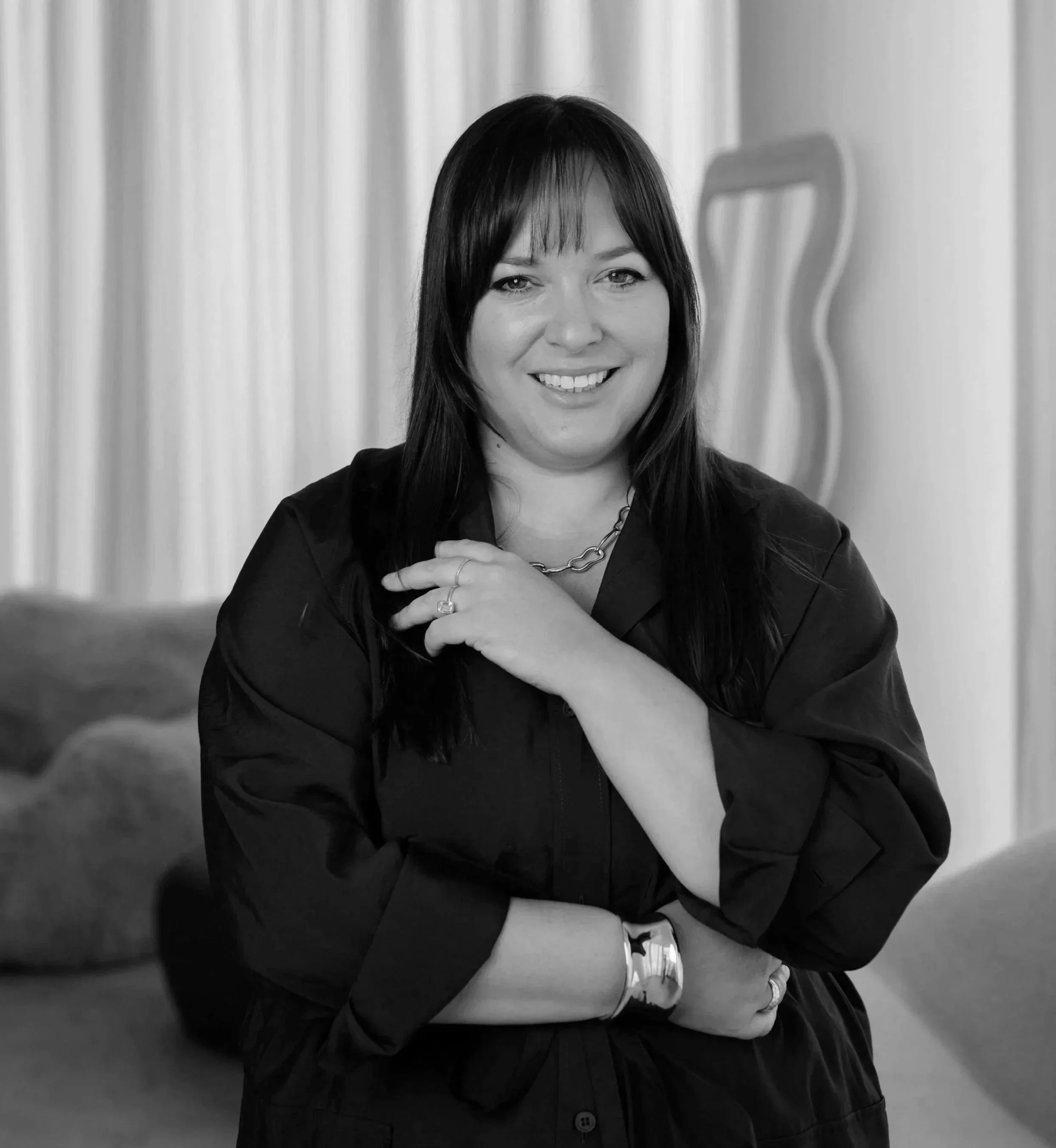 Black and white photo of a Laura Boatman with dark straight hair, smiling, wearing a dark blazer, a chain necklace, rings, and a watch, standing in front of curtains with a wavy sculpture in the background.