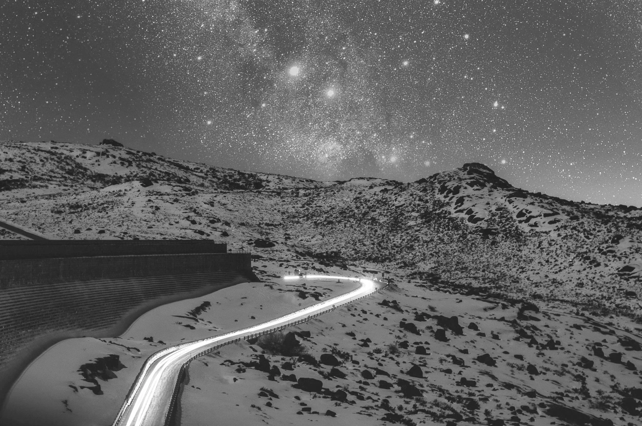 A black and white photo of a winding mountain road at night, with a star-filled sky and the Milky Way visible above.