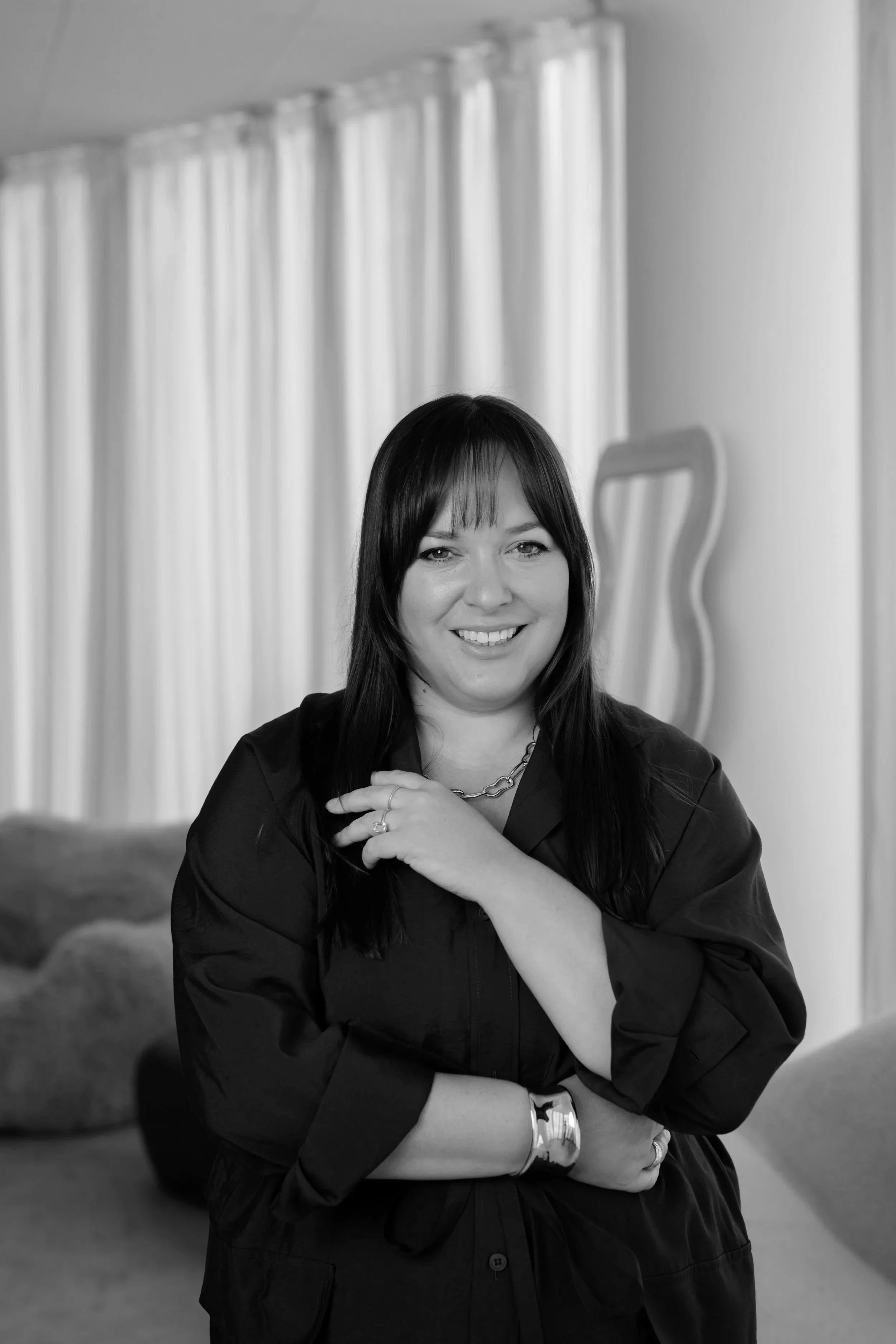Laura Boatman with dark hair and bangs smiling at the camera, standing indoors in front of a background with curtains and an artist's easel.