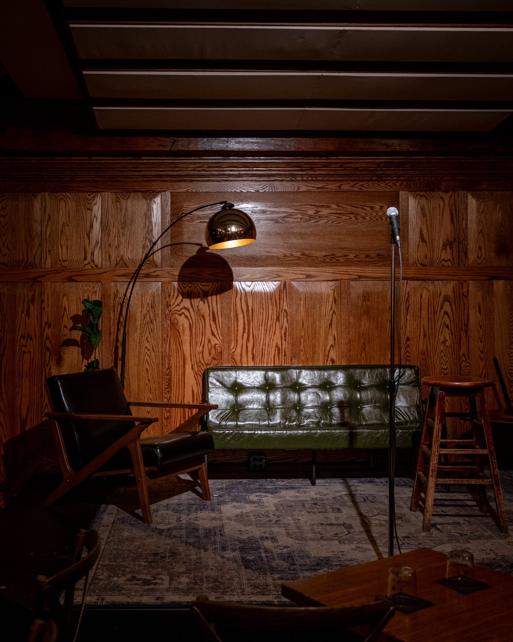 A dimly lit stage setup with a wooden stool, microphone on a stand, a leather chair, a green upholstered sofa, and a floor lamp, all set against a wooden paneled wall and a patterned rug.