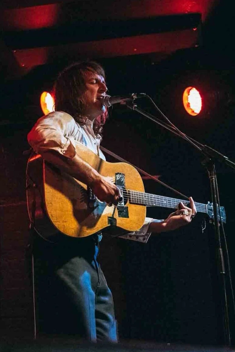 Scott klein musician performing on stage with an acoustic guitar, singing into a microphone, illuminated by red stage lights